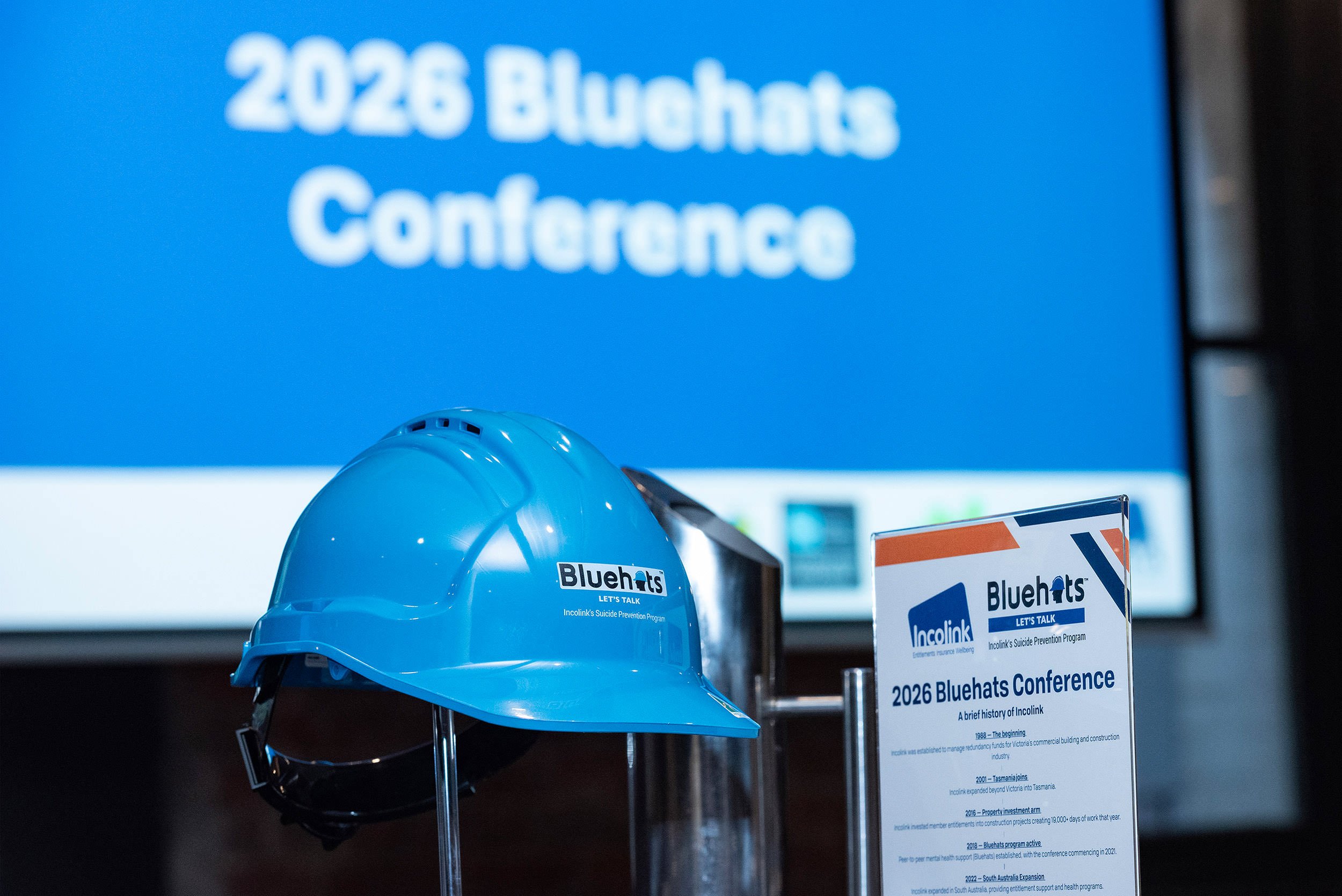 A blue Bluehats hard hat beside the printed conference programme, with the 2026 Bluehats Conference title on a screen behind it.