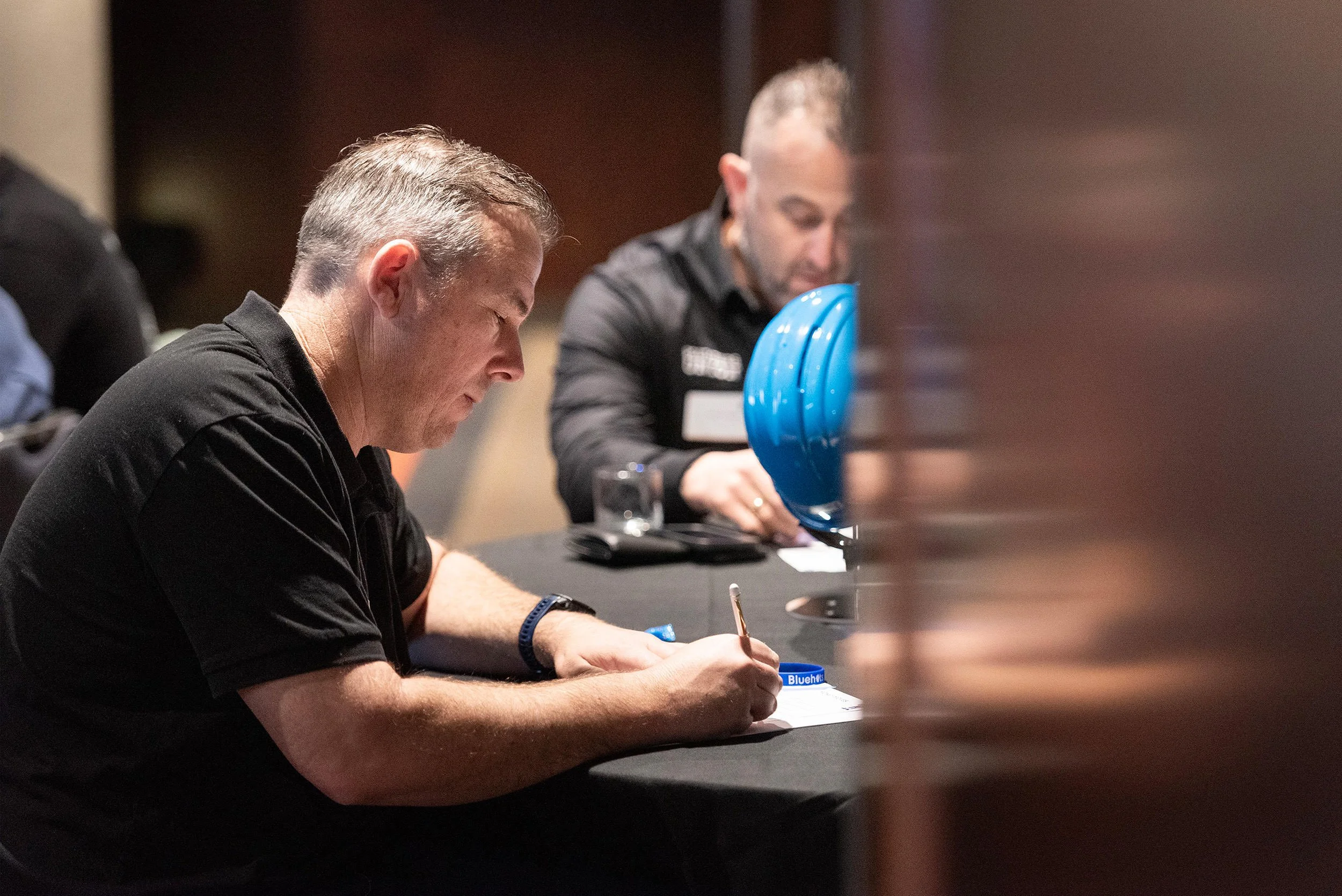 A seated delegate writes notes during the Bluehats Conference, with another attendee and a blue hard hat behind him.