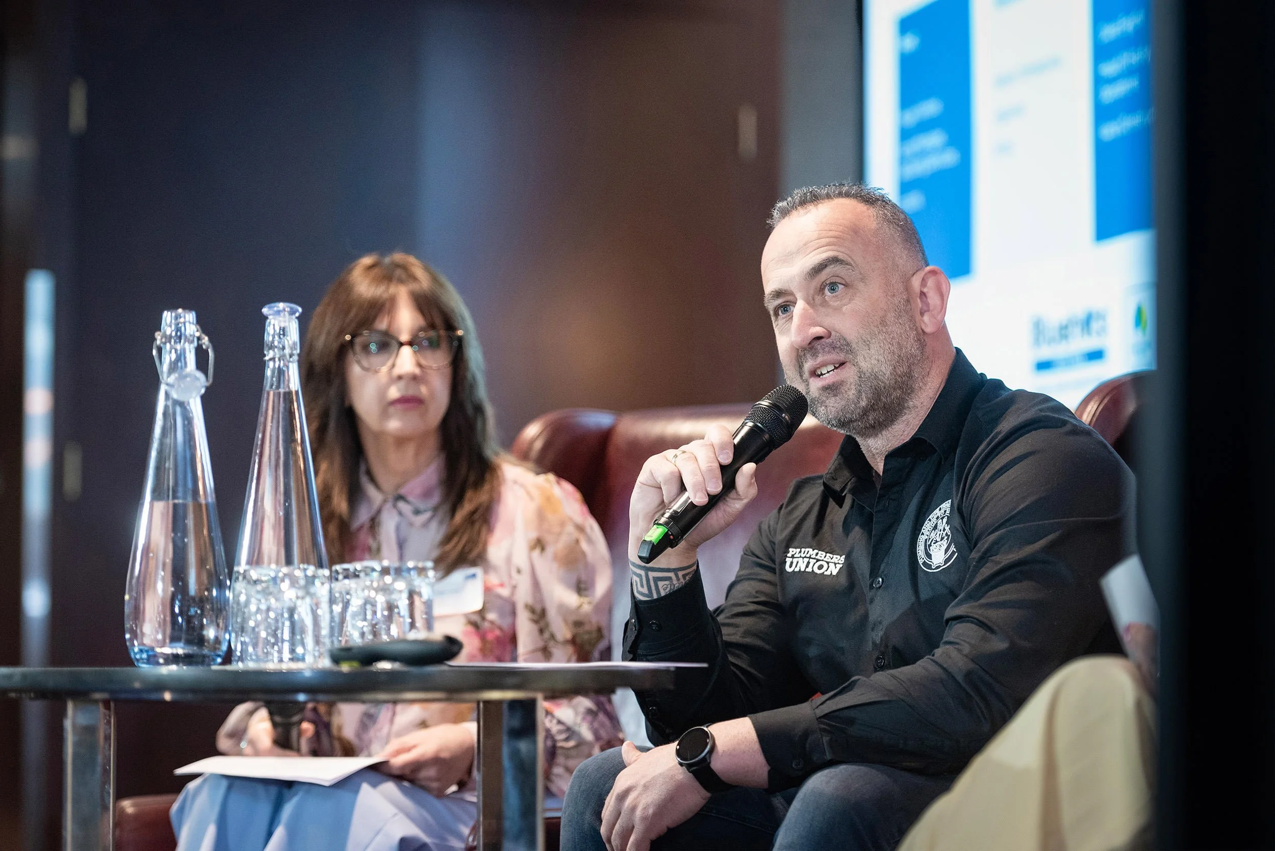 A seated man speaks into a handheld microphone while a fellow panellist listens beside him at the Bluehats Conference.