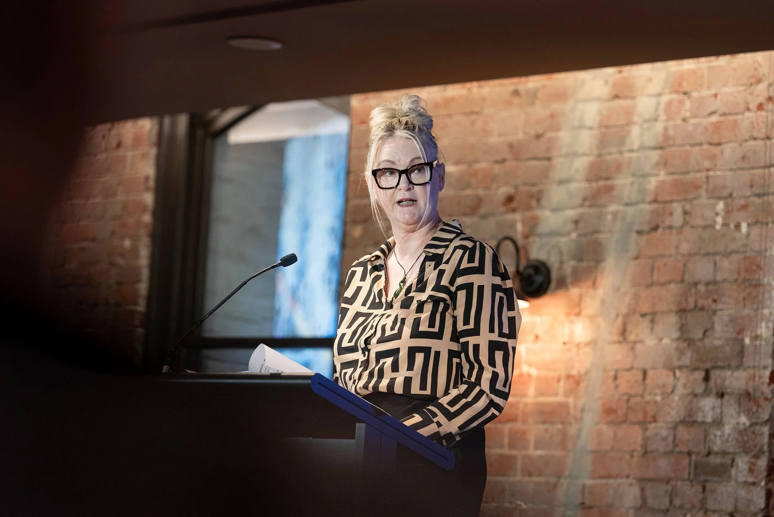 A woman wearing glasses and a patterned top speaks at a lectern, framed by a blurred foreground, at the Bluehats Conference.