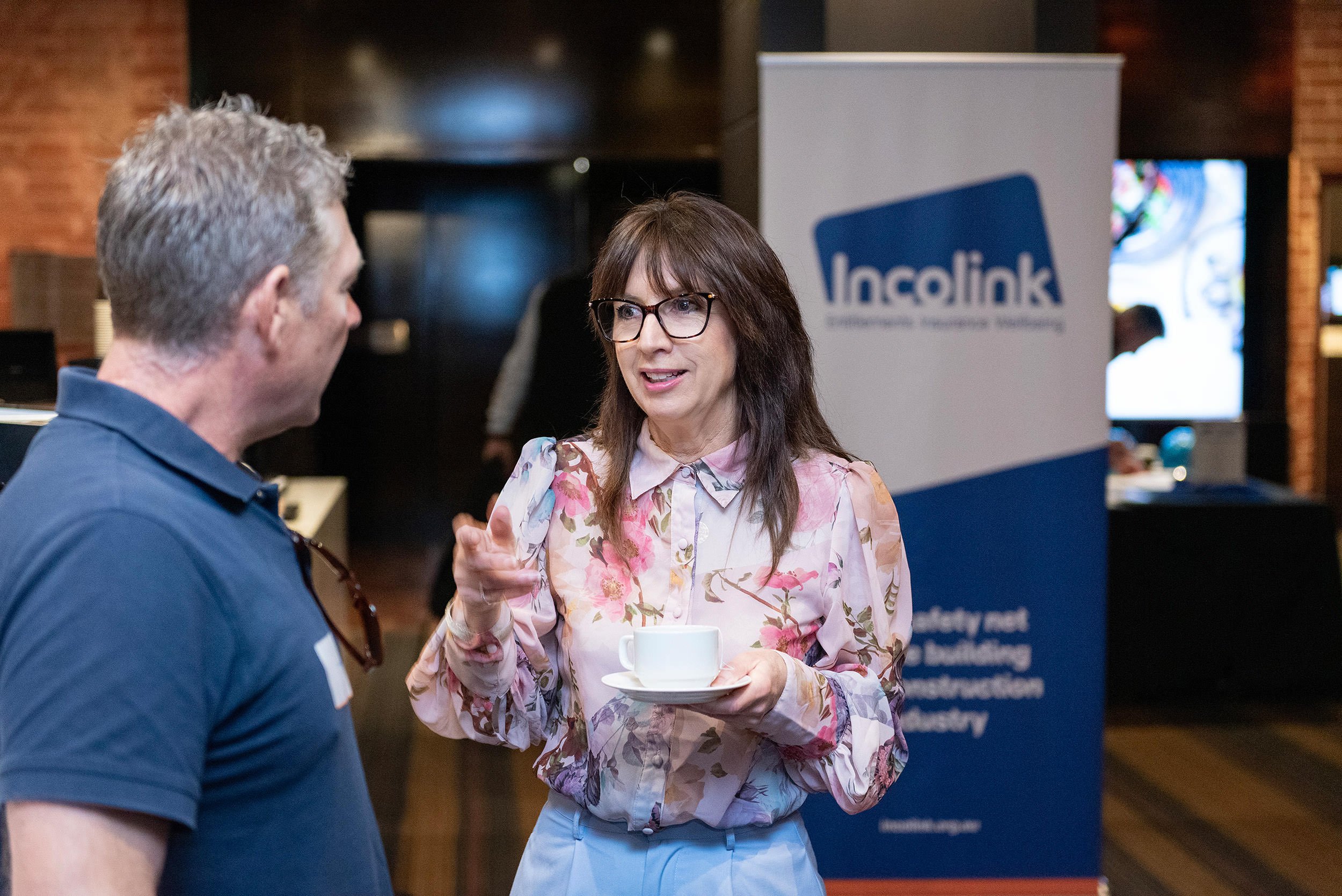 A woman holding a coffee cup gestures during a conversation with a delegate, beside an Incolink banner at the Bluehats Conference.