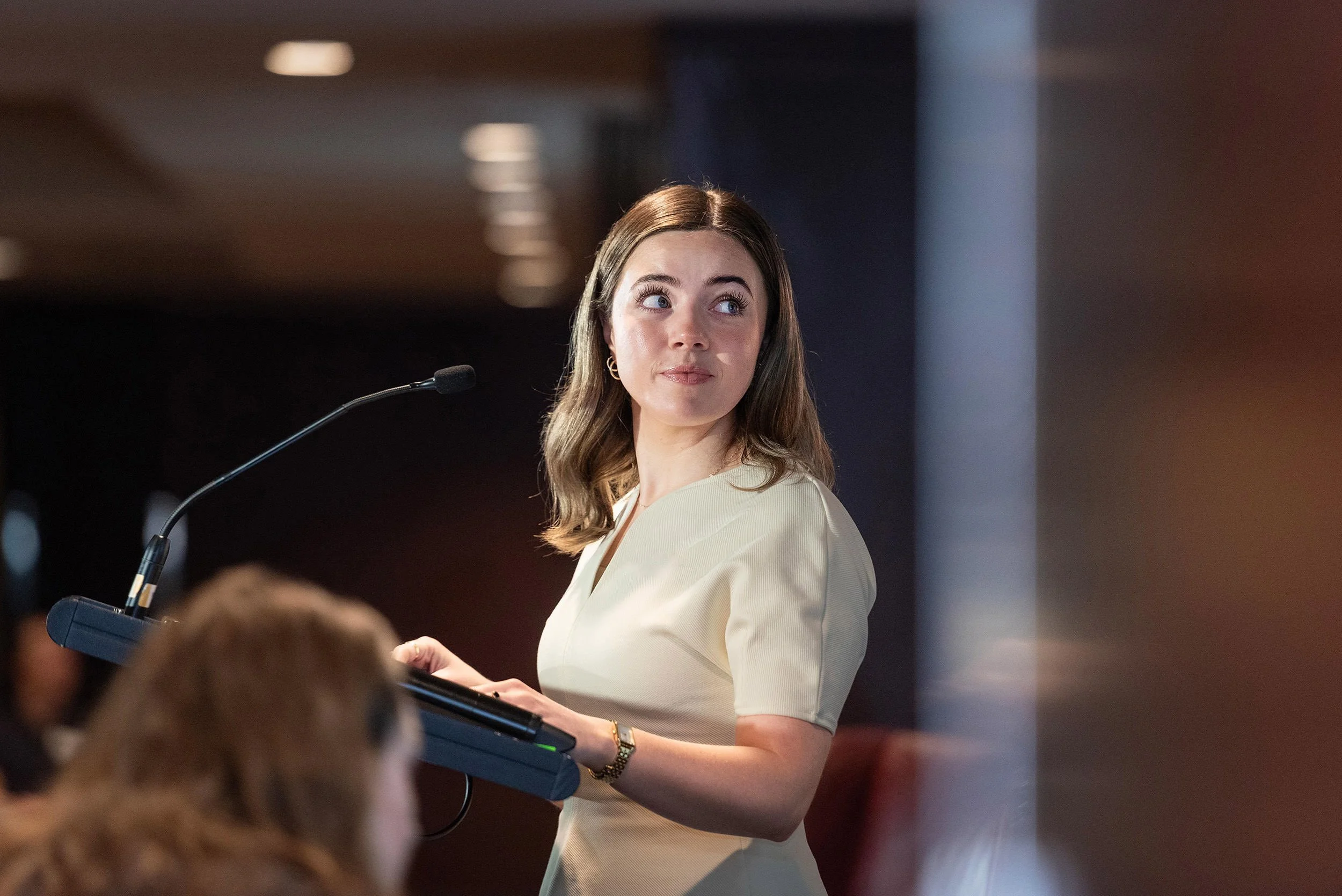 A woman in a cream shirt pauses at the lectern and looks across the room during the Bluehats Conference.