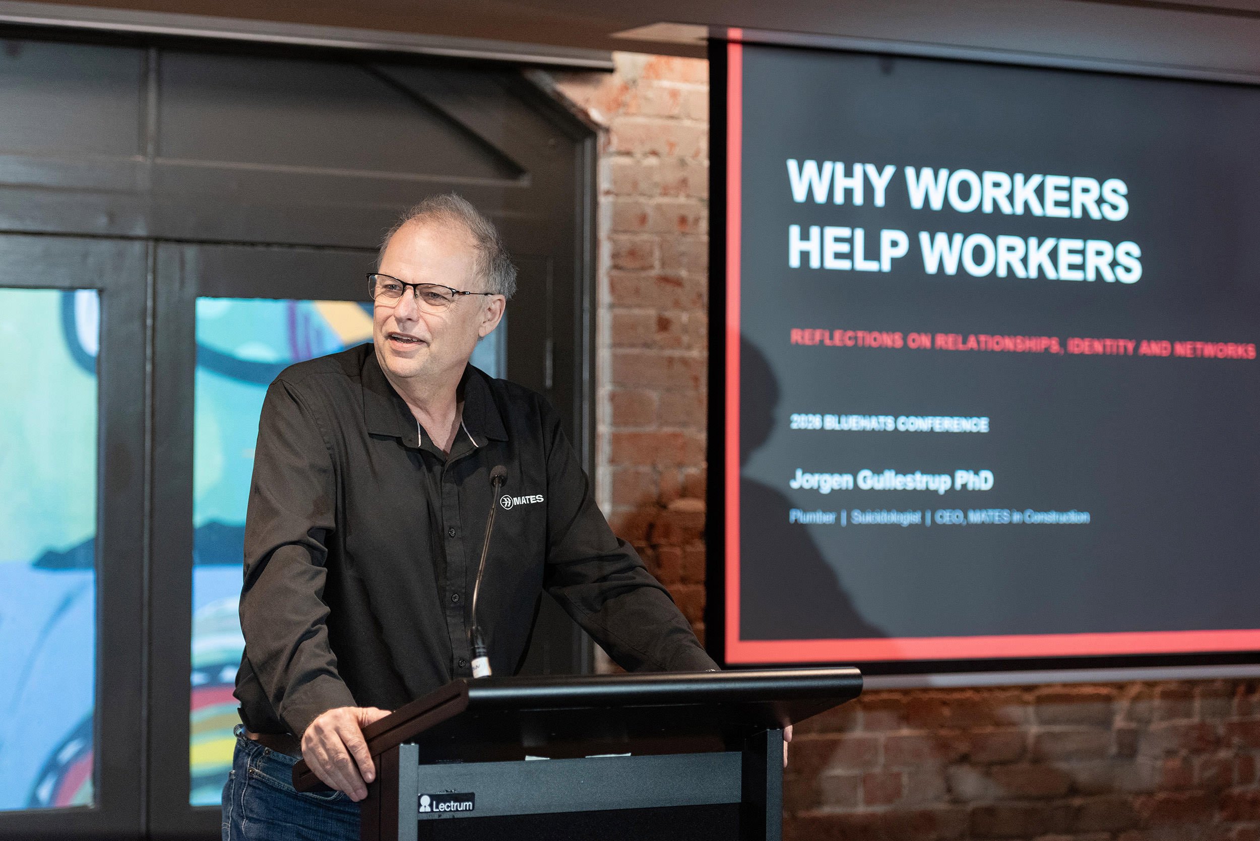 A man wearing glasses speaks at a lectern beside a presentation titled “Why Workers Help Workers” at the Bluehats Conference.