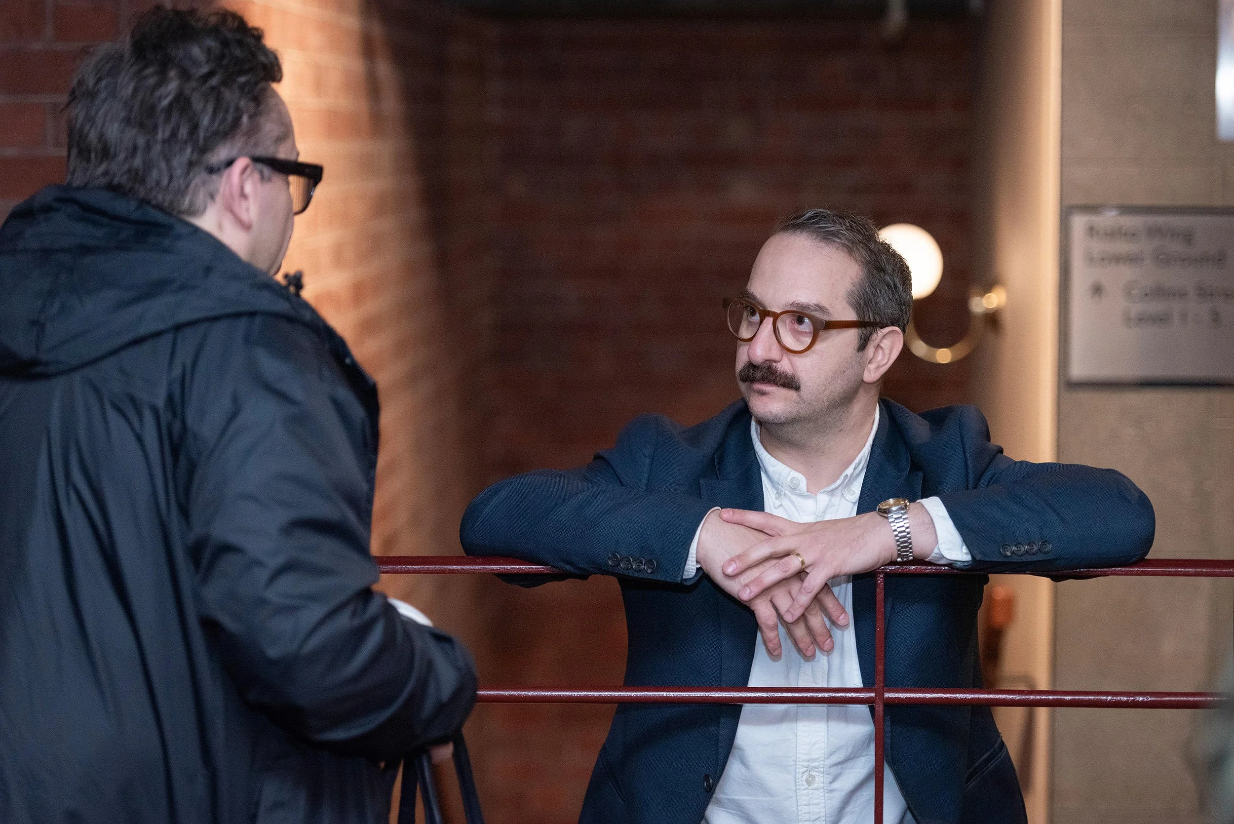 A delegate wearing glasses leans on a red railing while speaking with another attendee at the Bluehats Conference.