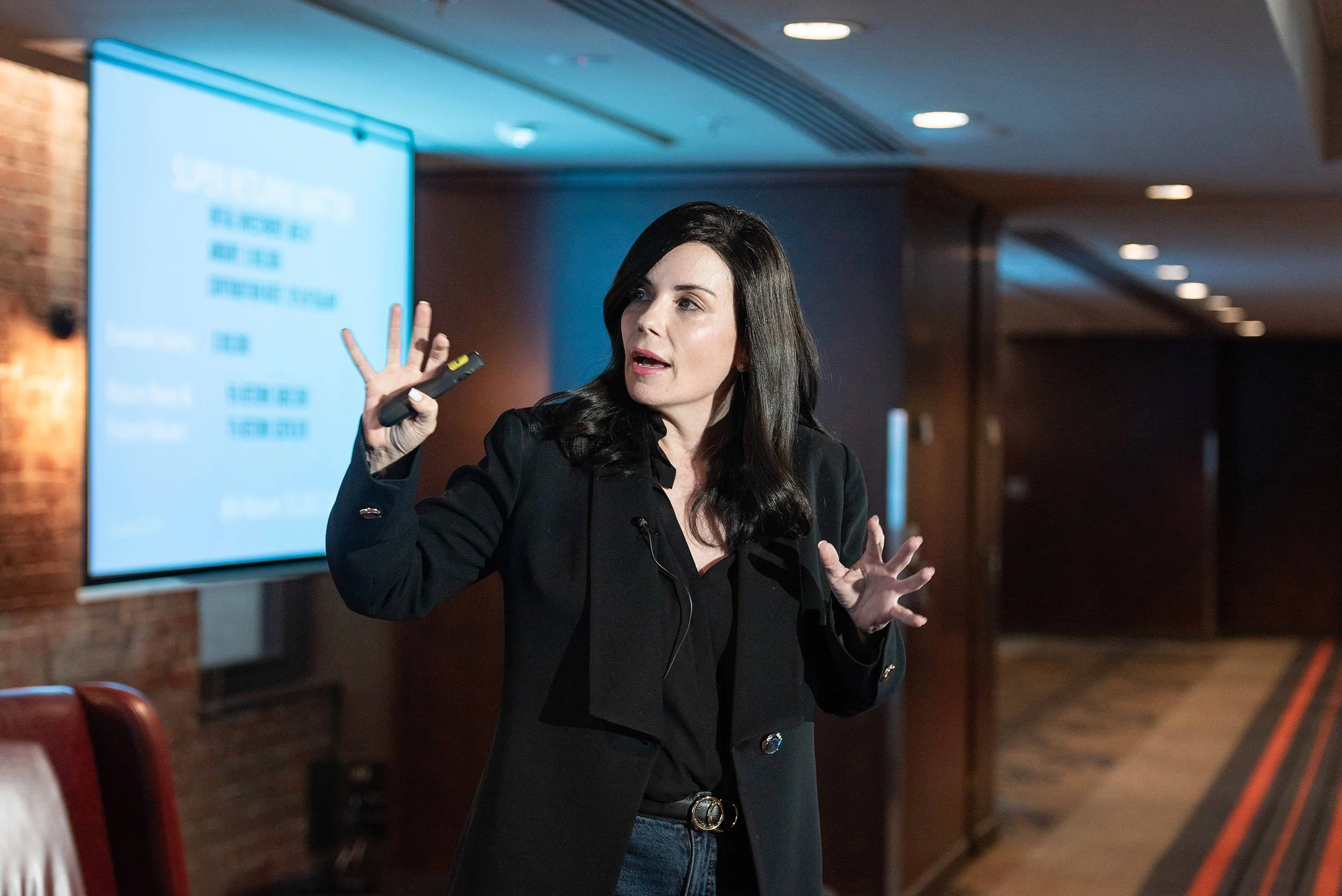 A dark-haired woman gestures with both hands while presenting beside a projection screen at the Bluehats Conference.