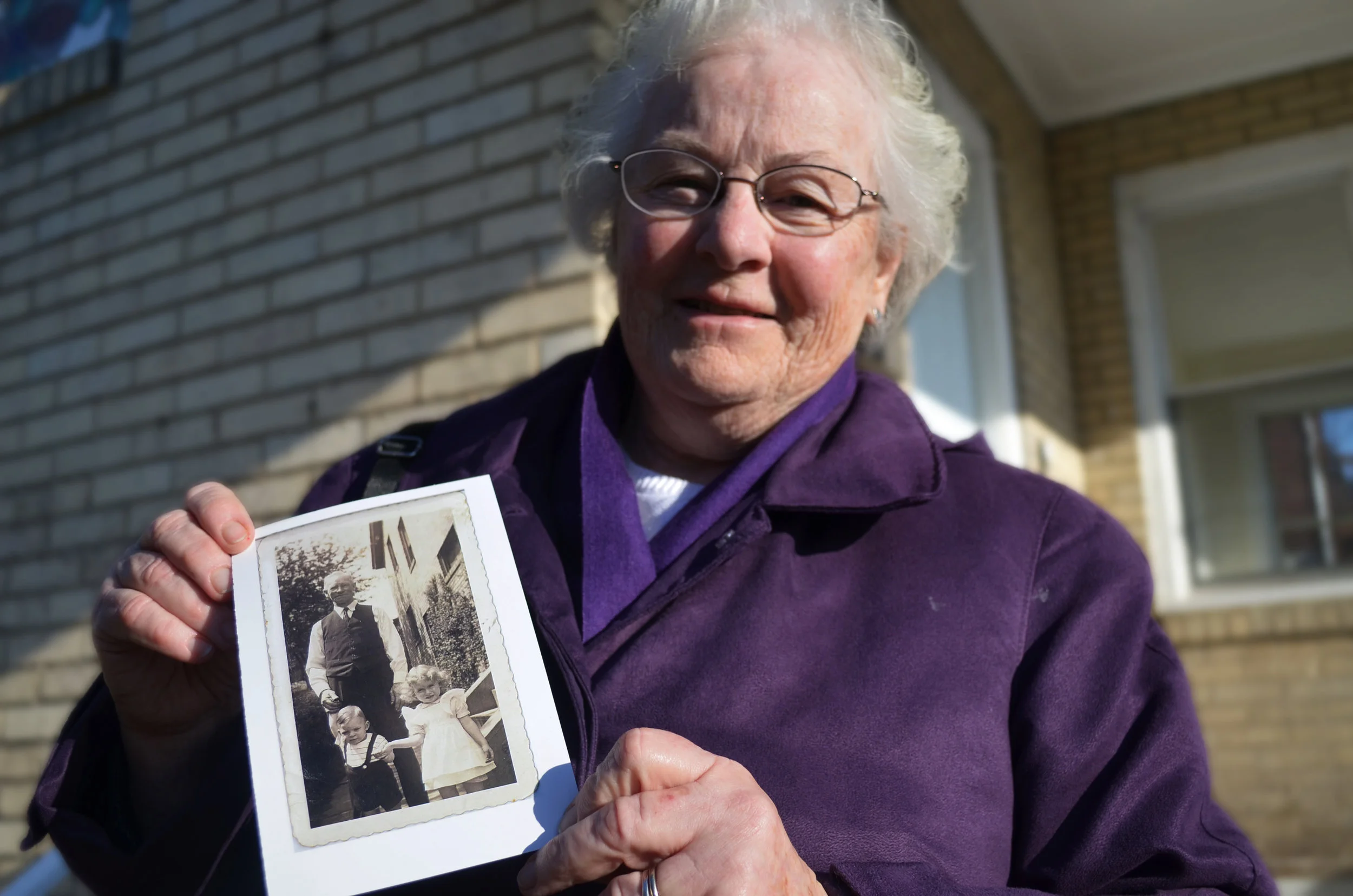 Sister Sheila holding a photograph featuring her grandfather, her brother, and herself in front of 3159 Ellers Street