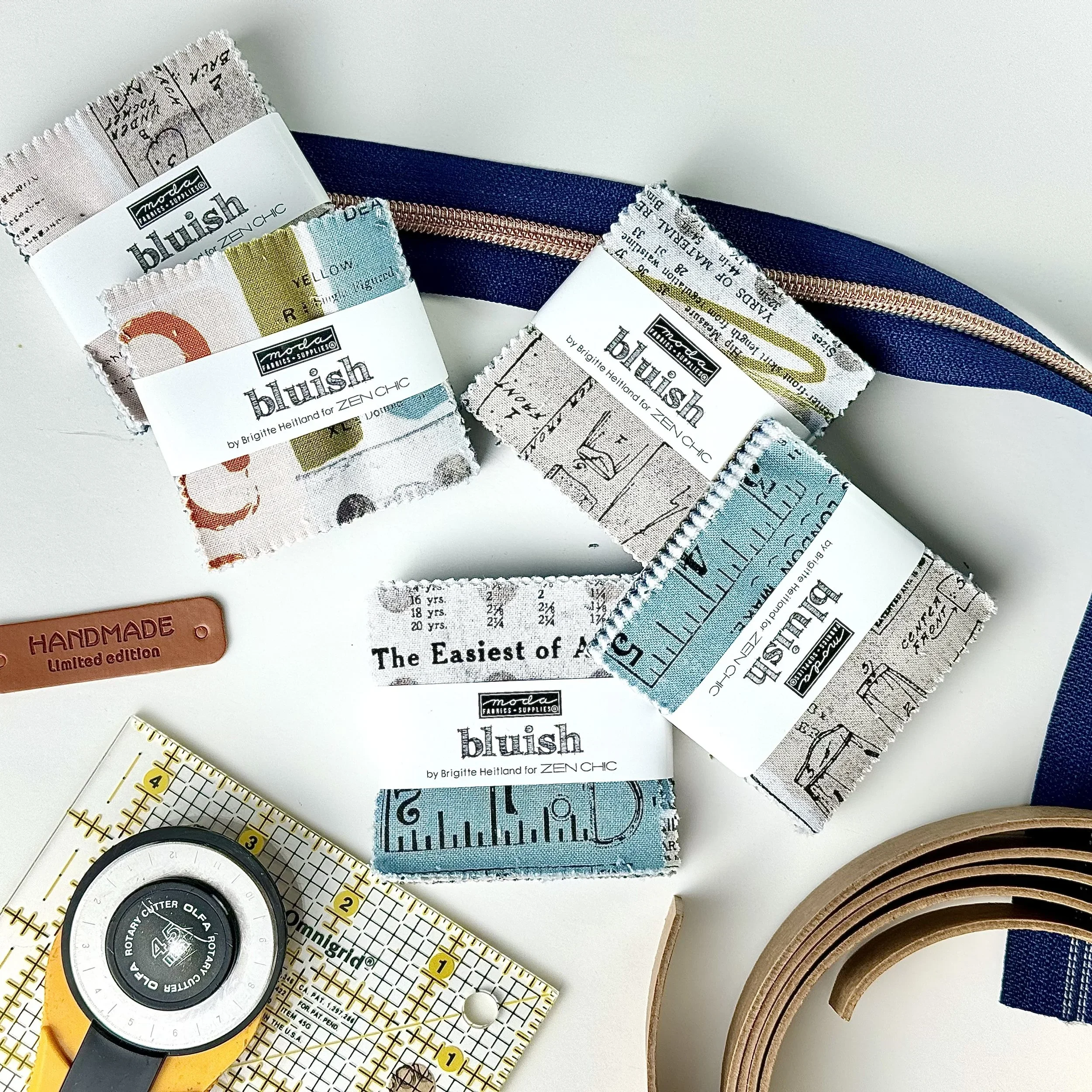Three Bluish Mini Charm Packs by Zen Chic lying on a white surface next to a zipper, with a handmade “Handmade, limited edition” label.