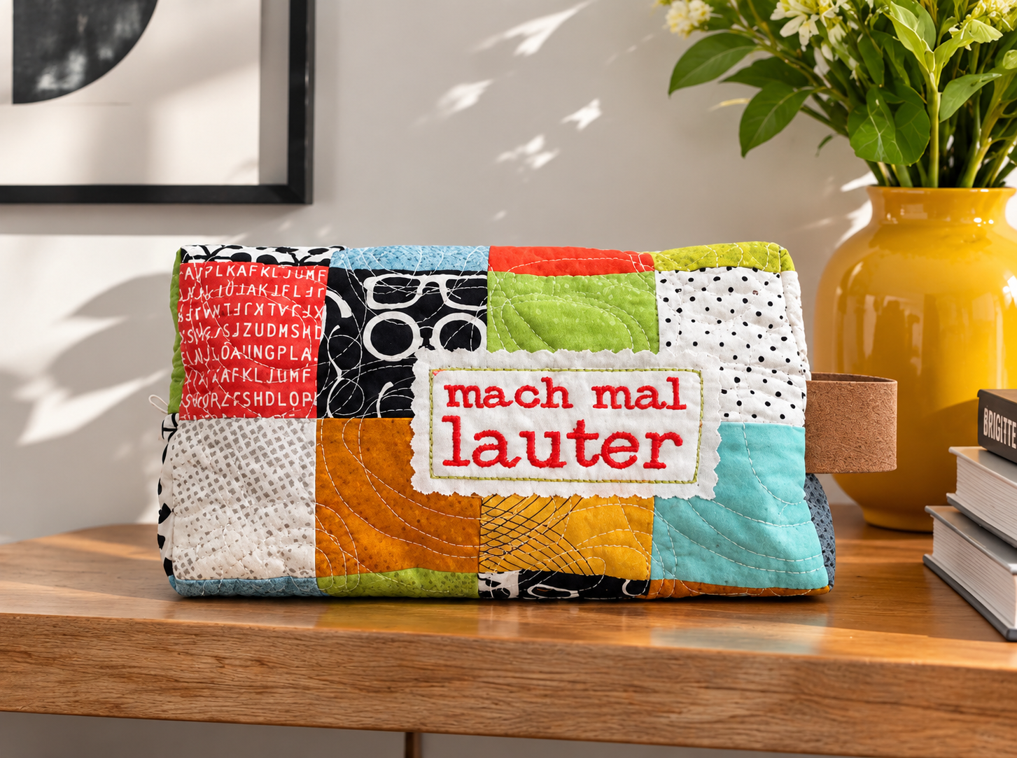 Quilted boxy pouch made from colourful 2.5-inch patchwork squares in red, black, green, orange and aqua, with an embroidered label reading “mach mal lauter”, on a wooden shelf next to a yellow vase and books.