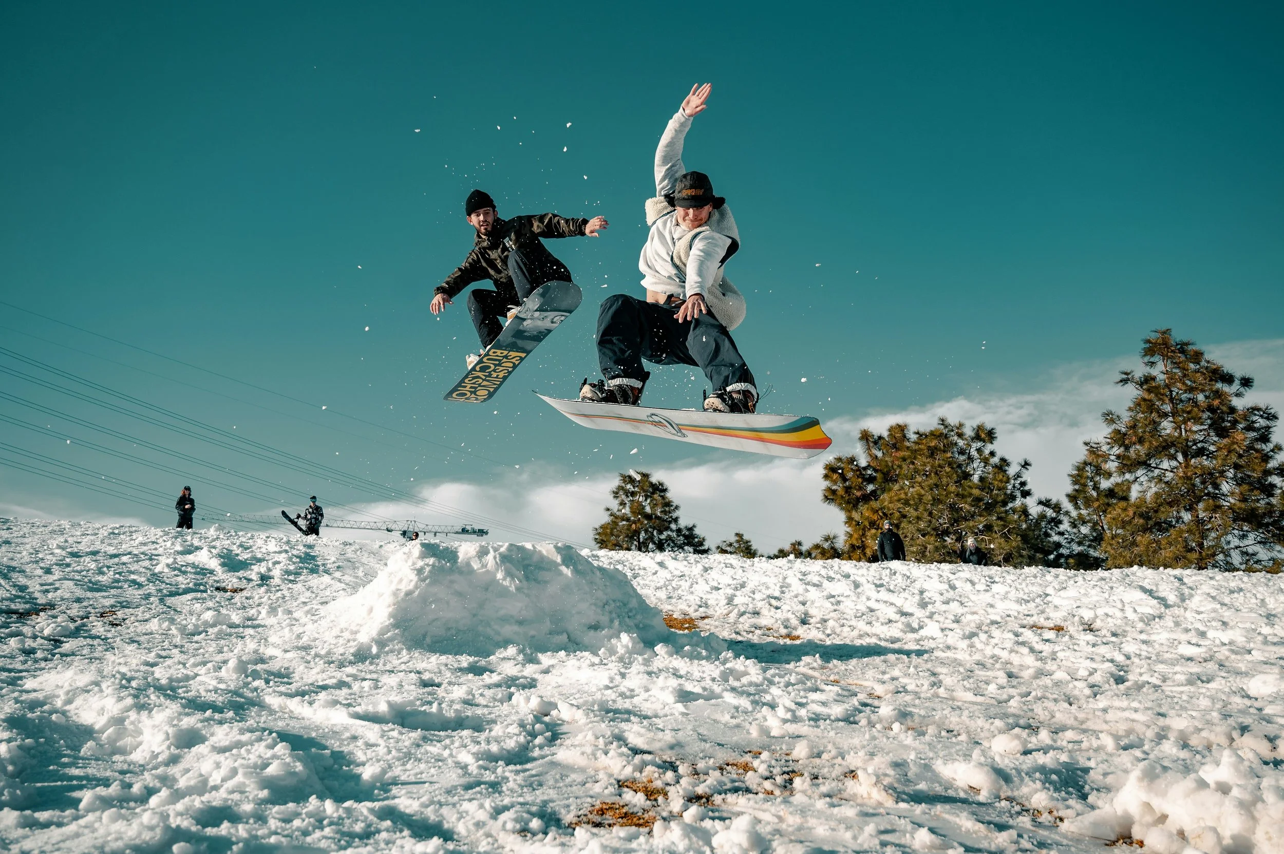 two people on snowboards jump through the air on a snowy hill, with blue sky in the background