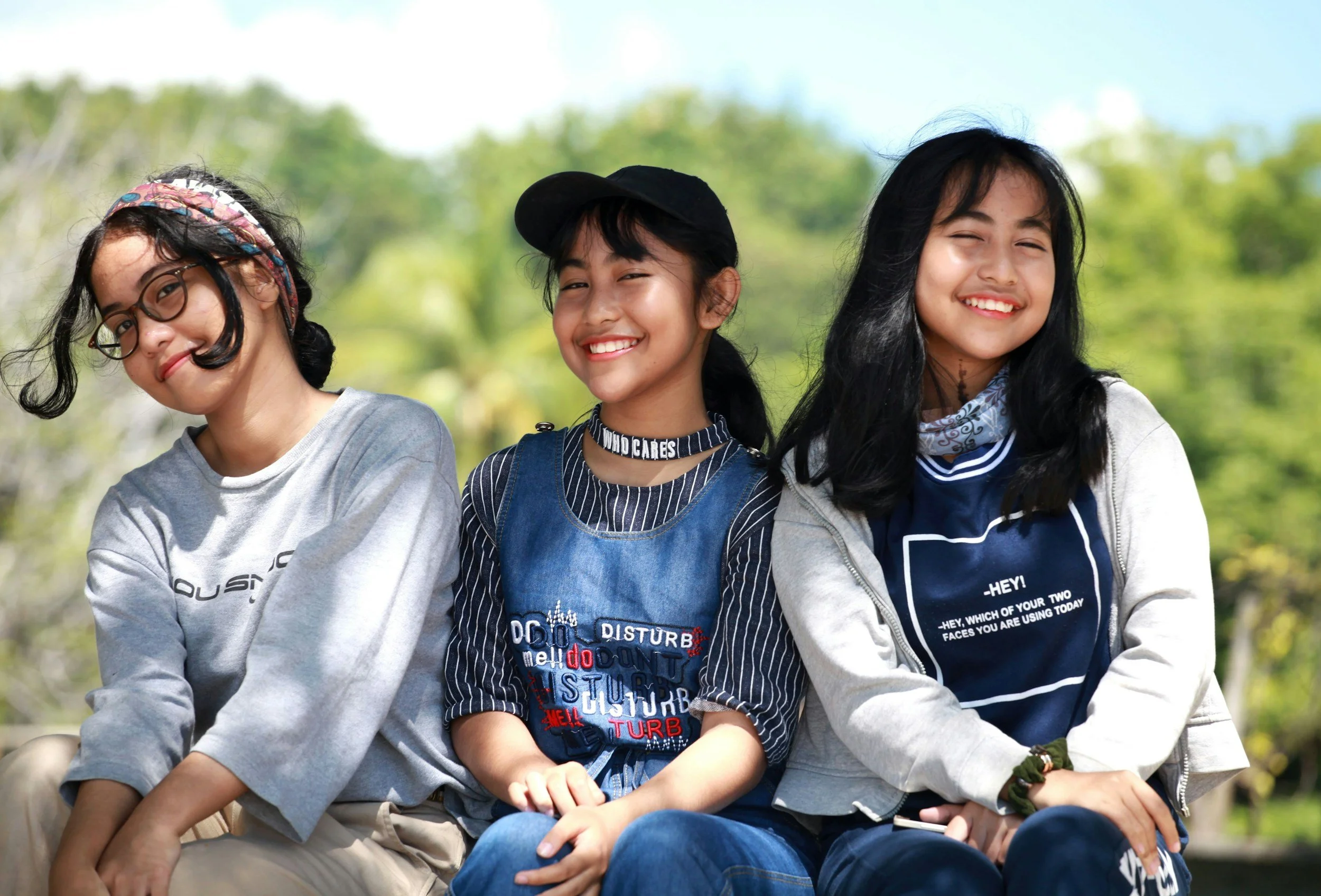 Three young people with long dark hair are sitting together and smiling. There are trees and blue sky in the background.