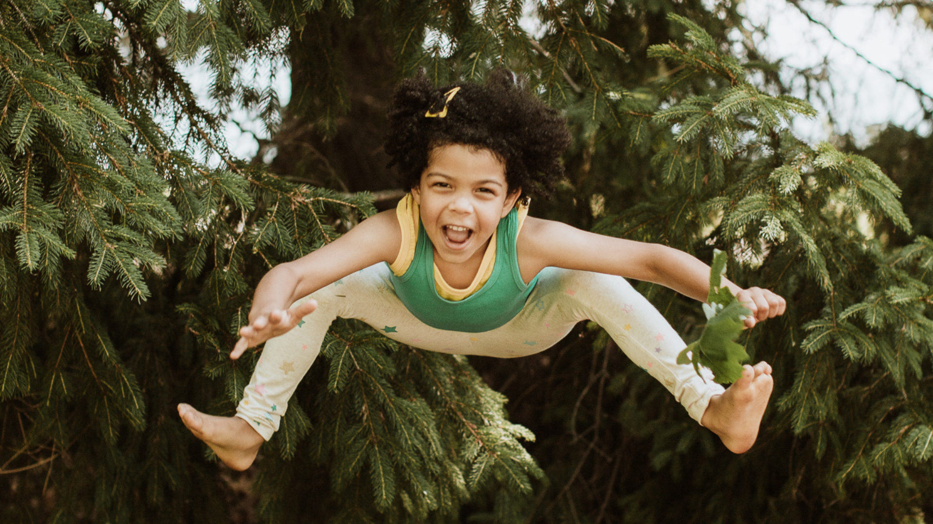 A child is jumping in mid-air with the arms and legs lifted. There are evergreen trees in the background.