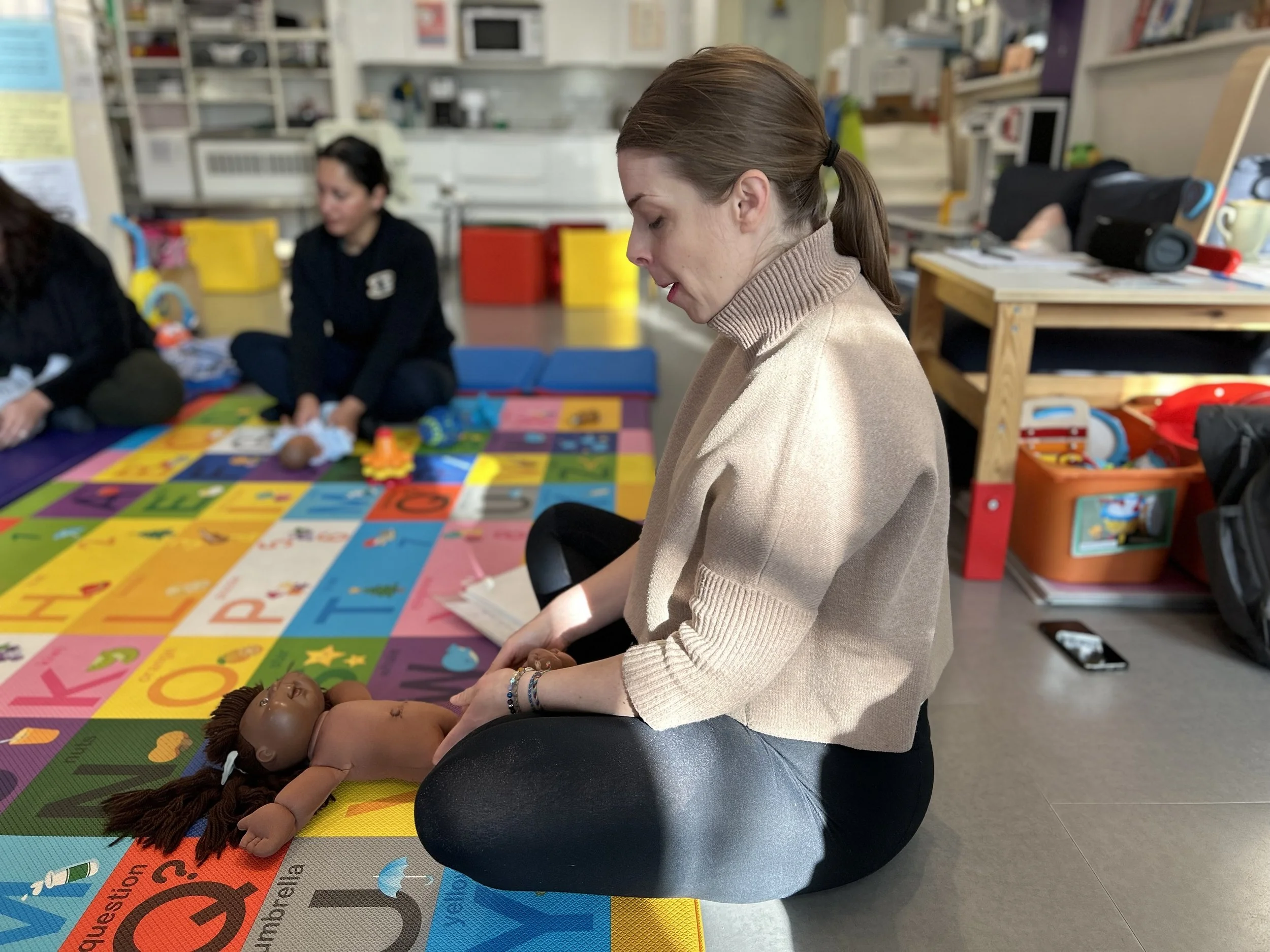 Megan sits cross-legged on a bright carpet, demonstrating baby yoga on a cabbage patch doll