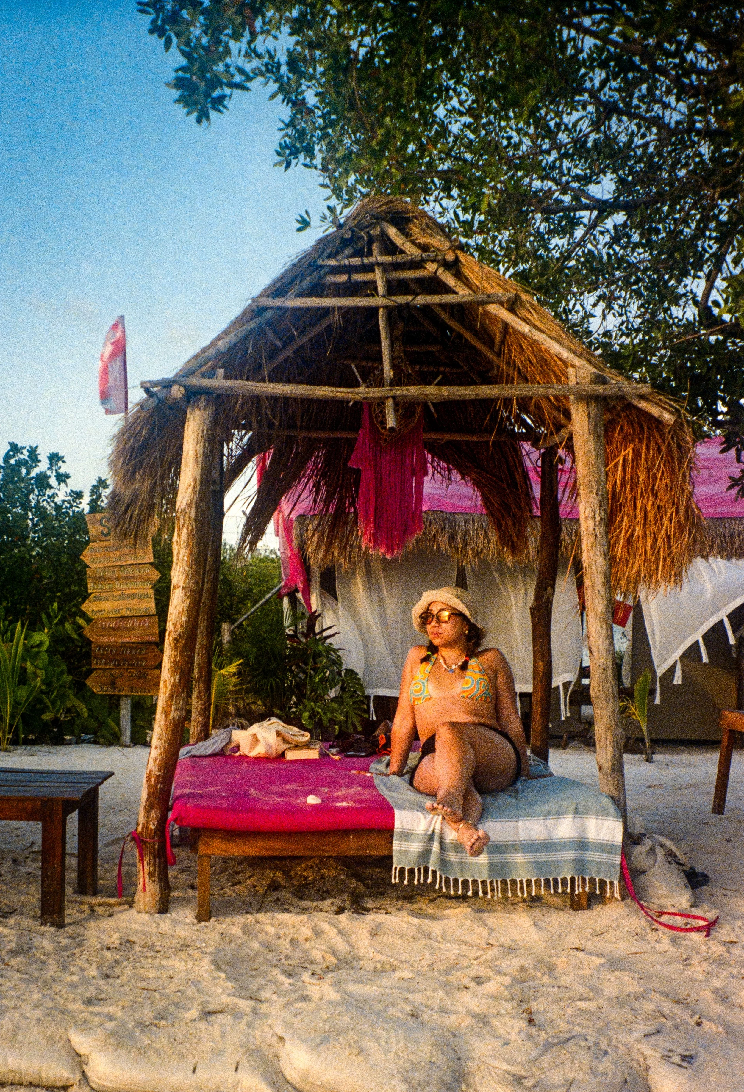 Woman sitting in a canopy made of wood and straw on a sandy beach, wearing sunglasses, a hat, and a swimsuit with a colorful pattern, surrounded by trees and beach furniture.
