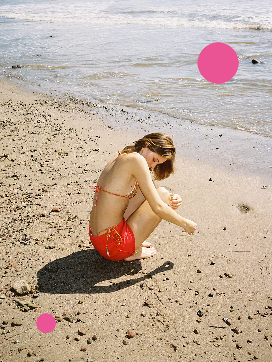A woman with light skin and brown hair sitting on a sandy beach, wearing a colorful bikini top and red shorts, drawing in the sand with her finger near the shoreline. The ocean and some rocks are visible in the background.