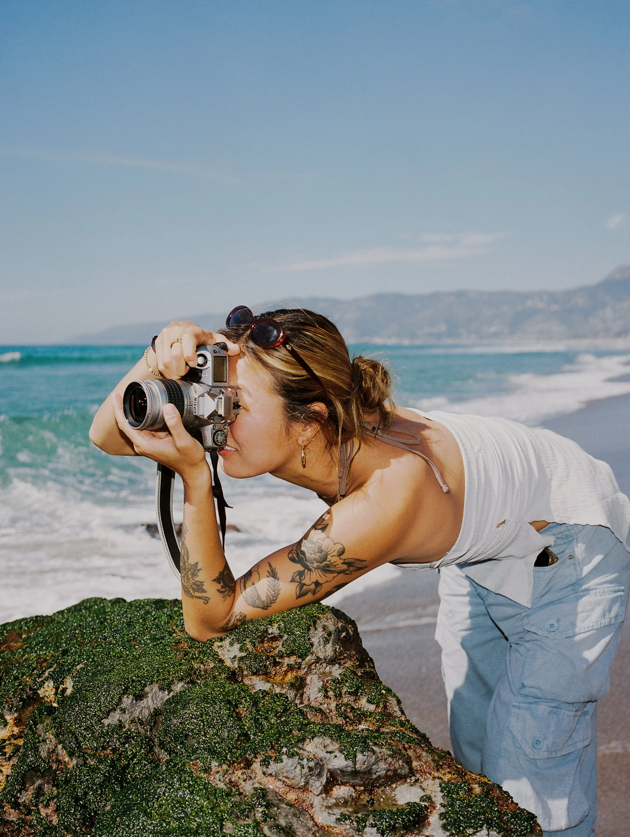 Woman with tattoos taking a photo at the beach, leaning on a moss-covered rock with the ocean and mountains in the background.