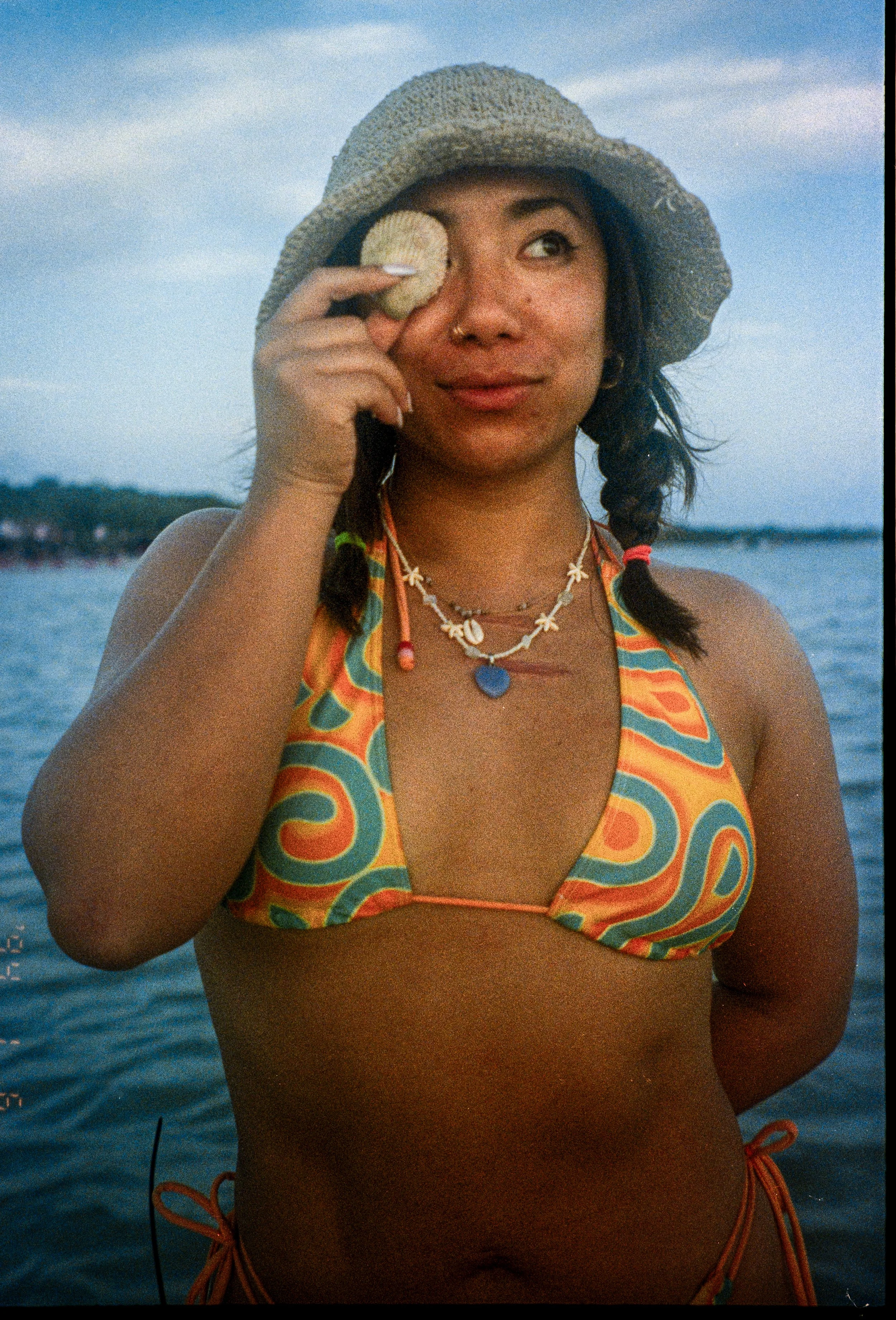 A woman in a colorful swimsuit holding a shell to her eye while standing by a body of water during sunset.