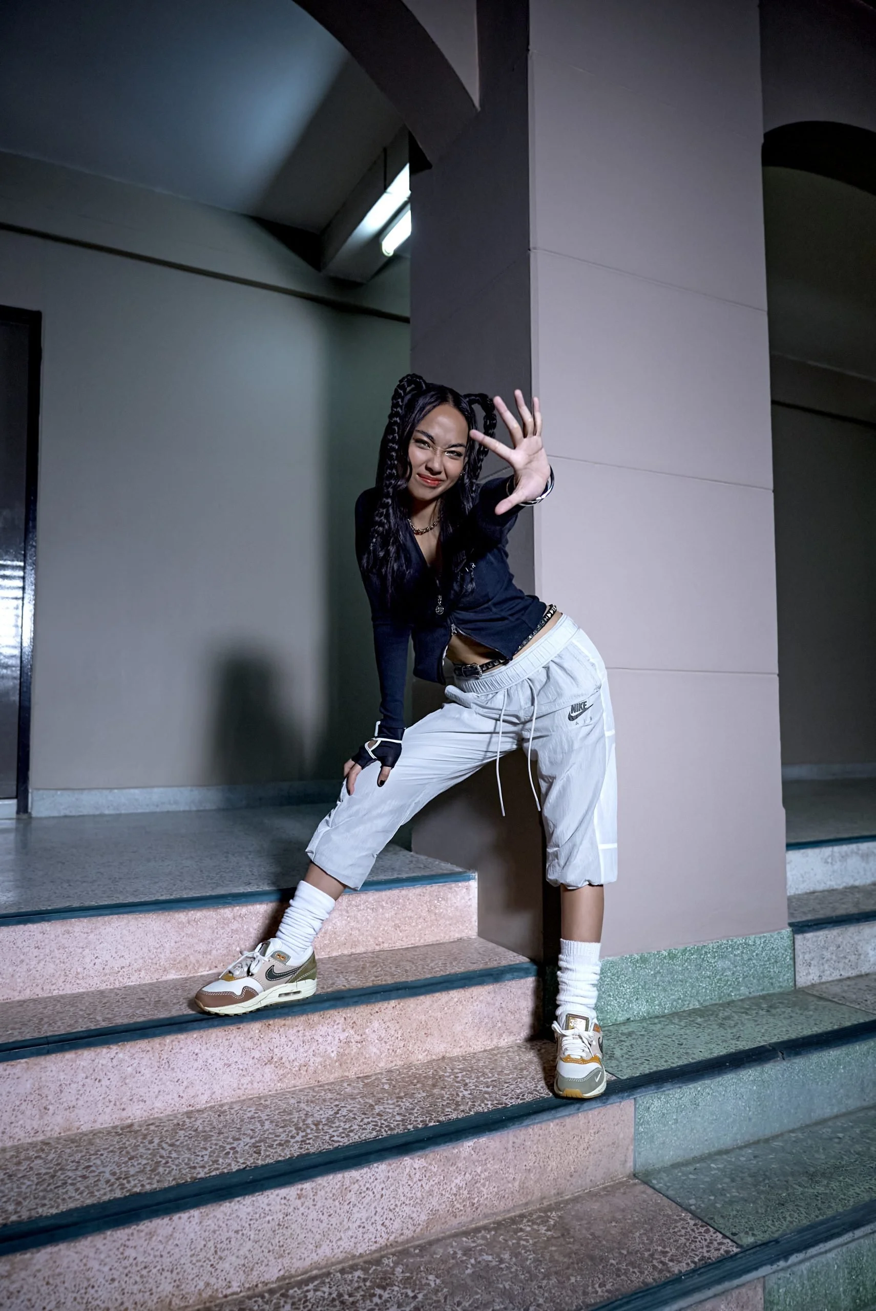 A young woman with braided hair posing on stairs in an indoor setting, wearing a black top, beige Nike pants, and sneakers, with her hand extended toward the camera.