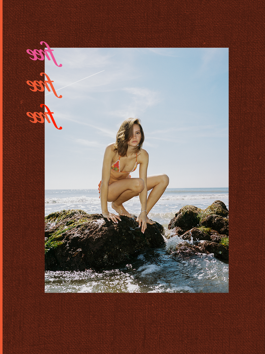 A woman in a bikini crouching on rocks by the ocean under a blue sky.