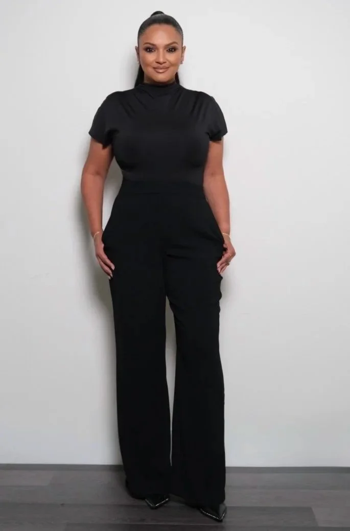 A woman with dark hair pulled back, wearing a black short-sleeved turtleneck top and black pants standing against a plain white wall.
