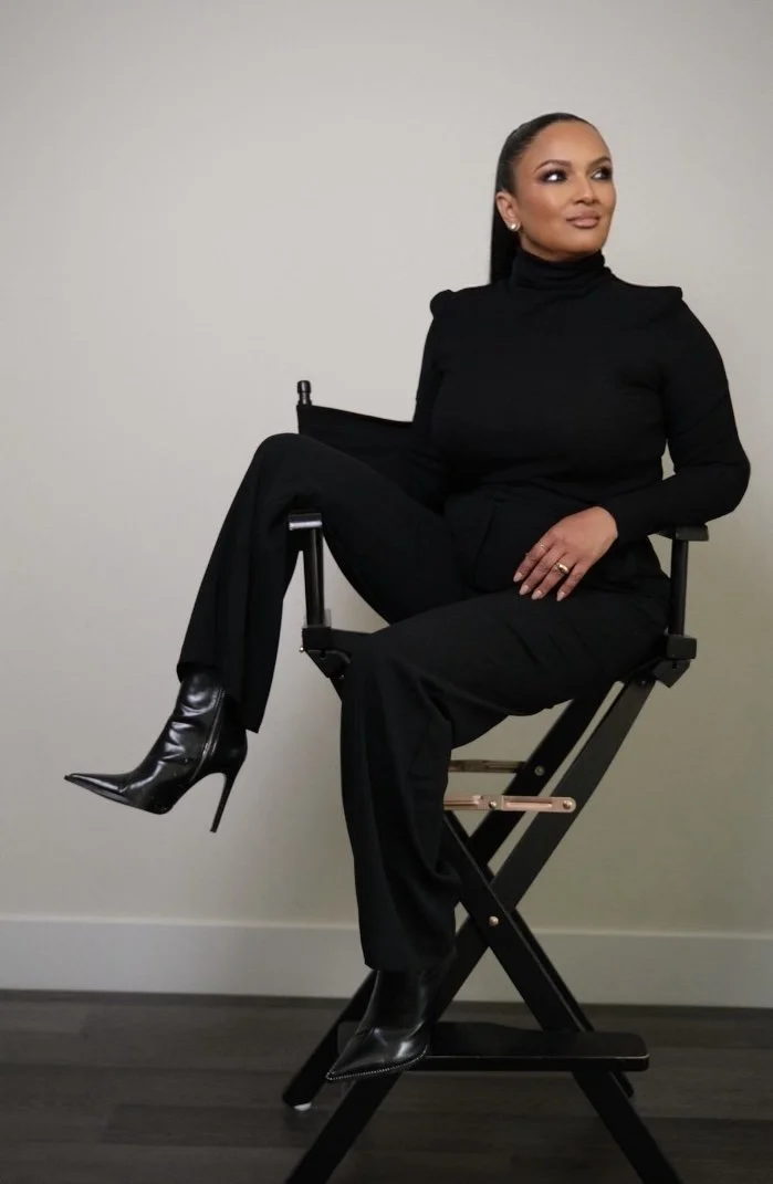 A Makeup Artist in NYC  in black turtleneck and pants, sitting on a director's chair, wearing black high-heeled boots with a pointed toe, posing against a plain light-colored wall.