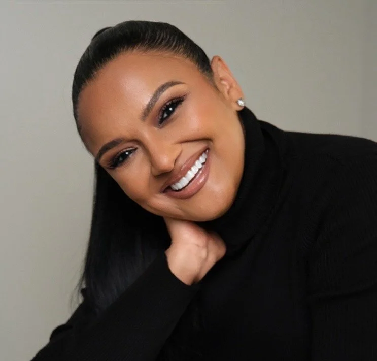 Smiling woman with dark hair styled in a high ponytail, wearing pearl earrings and a black turtleneck, resting her chin on her hand against a plain background.