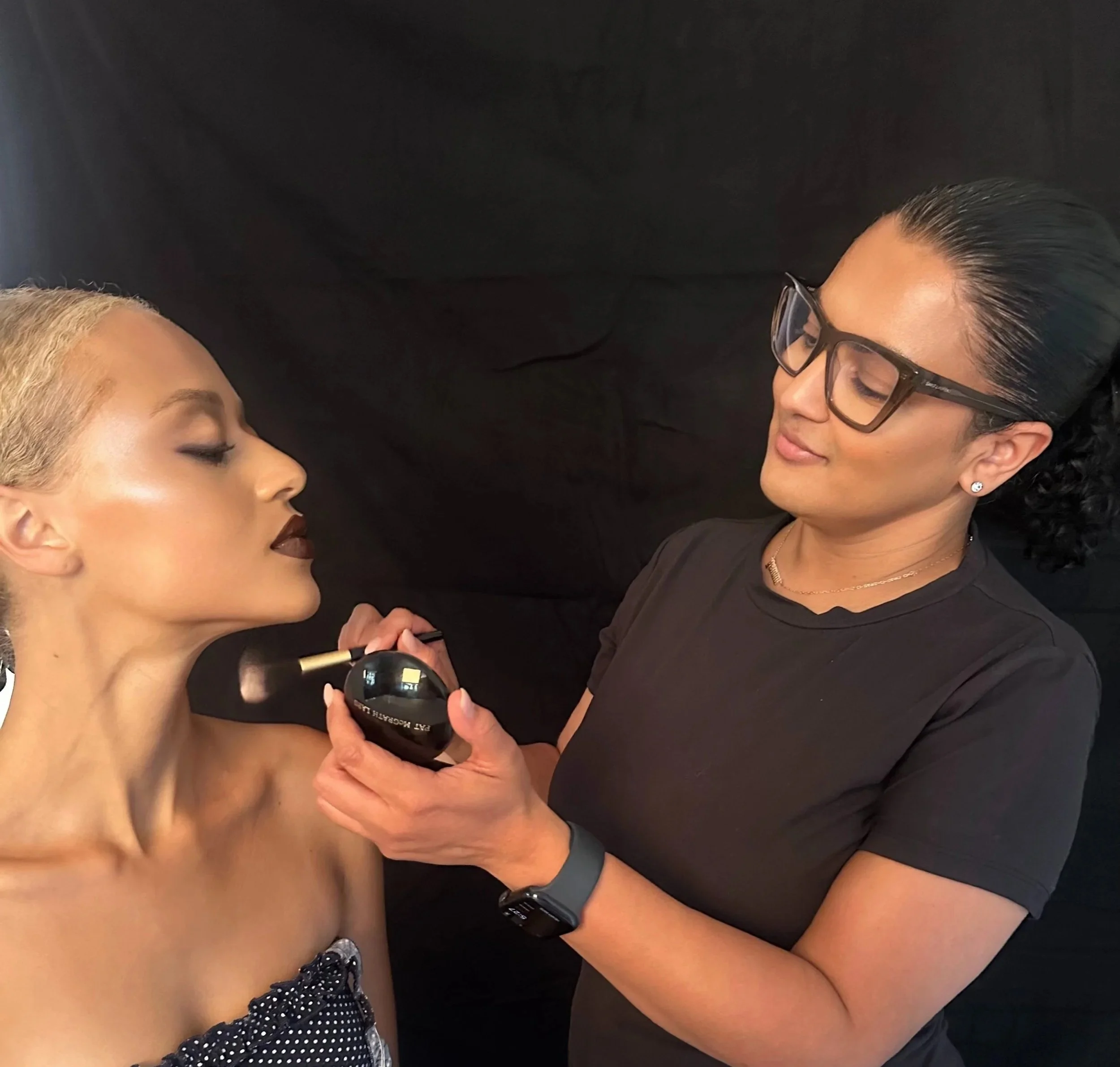 A makeup artist in NYC applying makeup to a woman's face while the woman has her eyes closed against a black background.