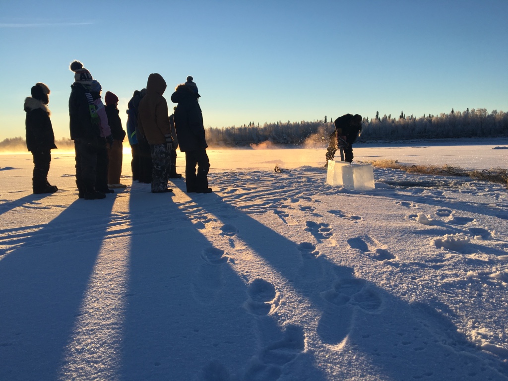 Huslia JHS students observe how to cut holes in ice to set fish net.