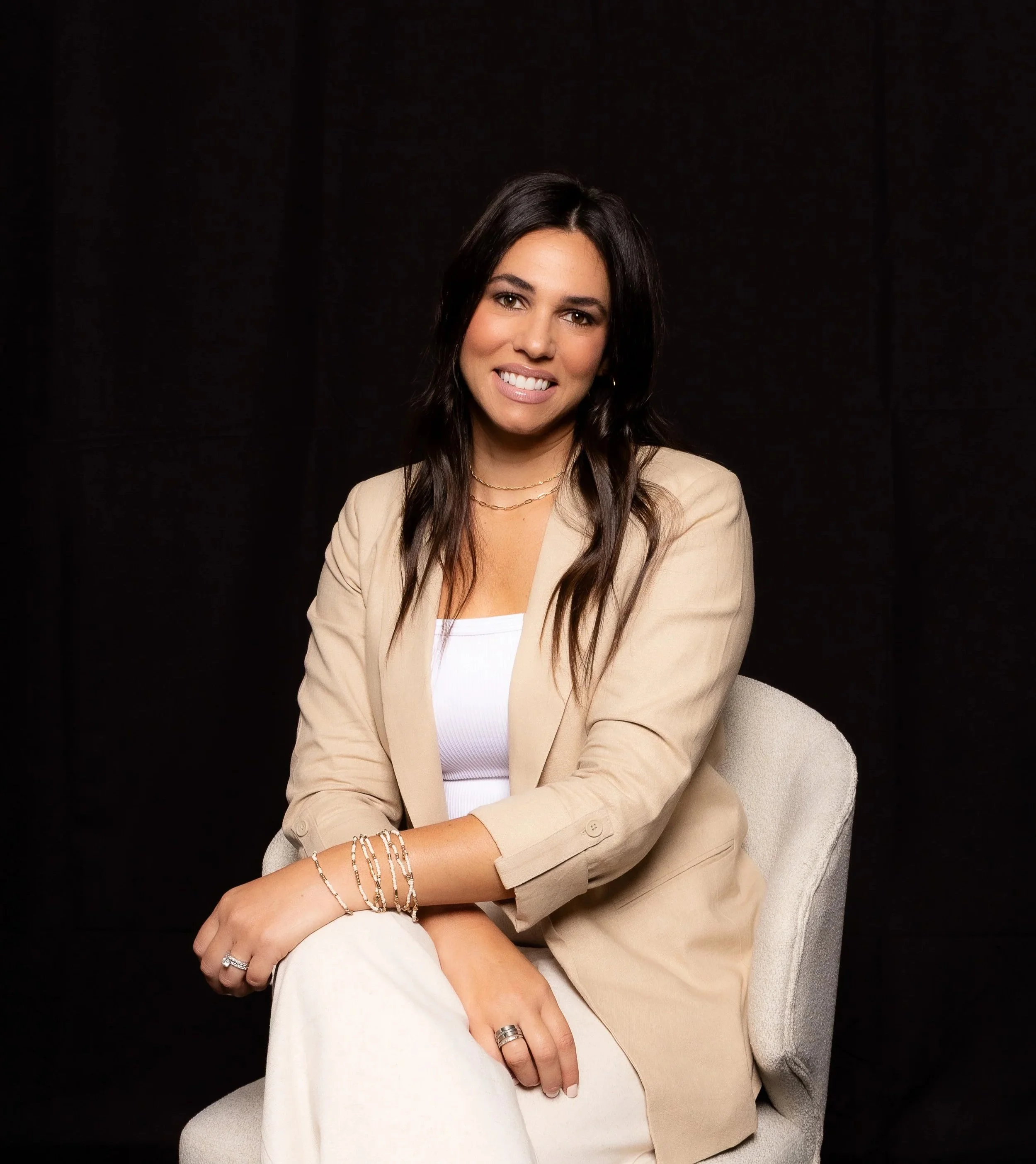A woman with long dark hair smiling at the camera, sitting on a light-colored chair, wearing a beige blazer, white top, and bracelets, against a dark background.