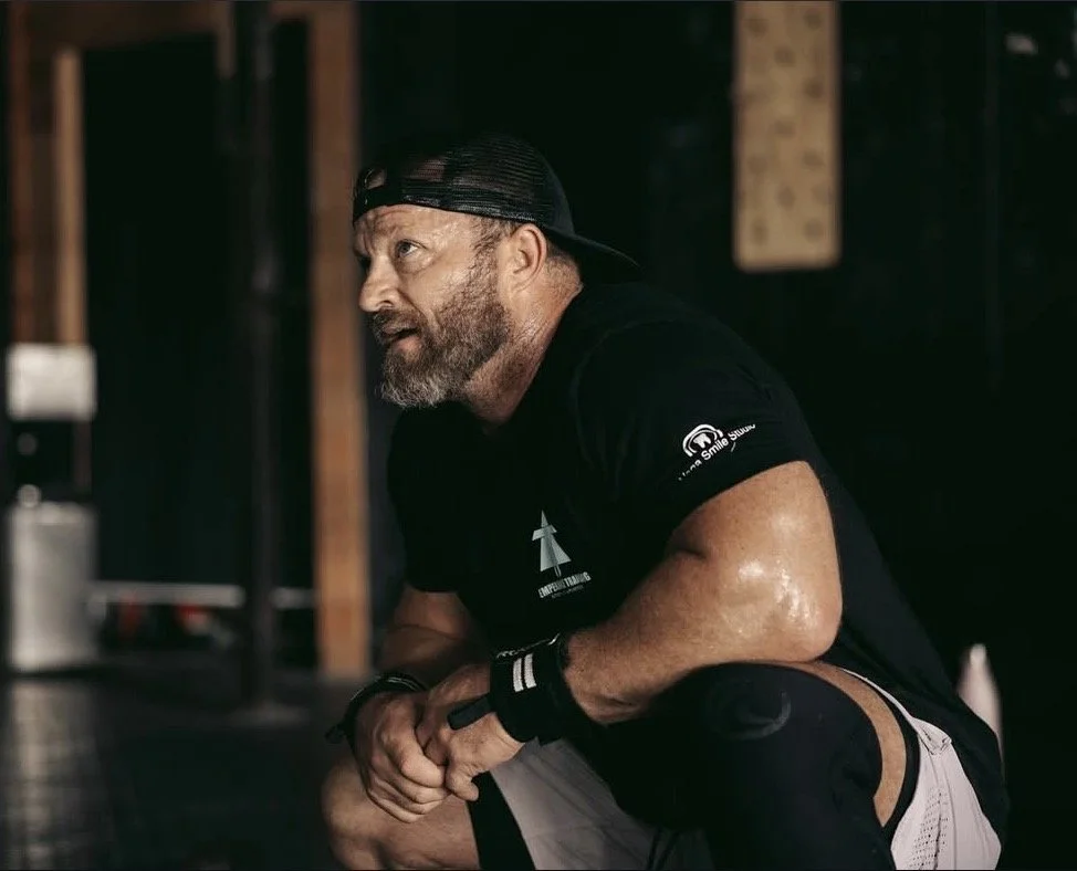 A man wearing a black T-shirt, black cap backward, and workout gloves, sitting on a gym floor with a focused expression.