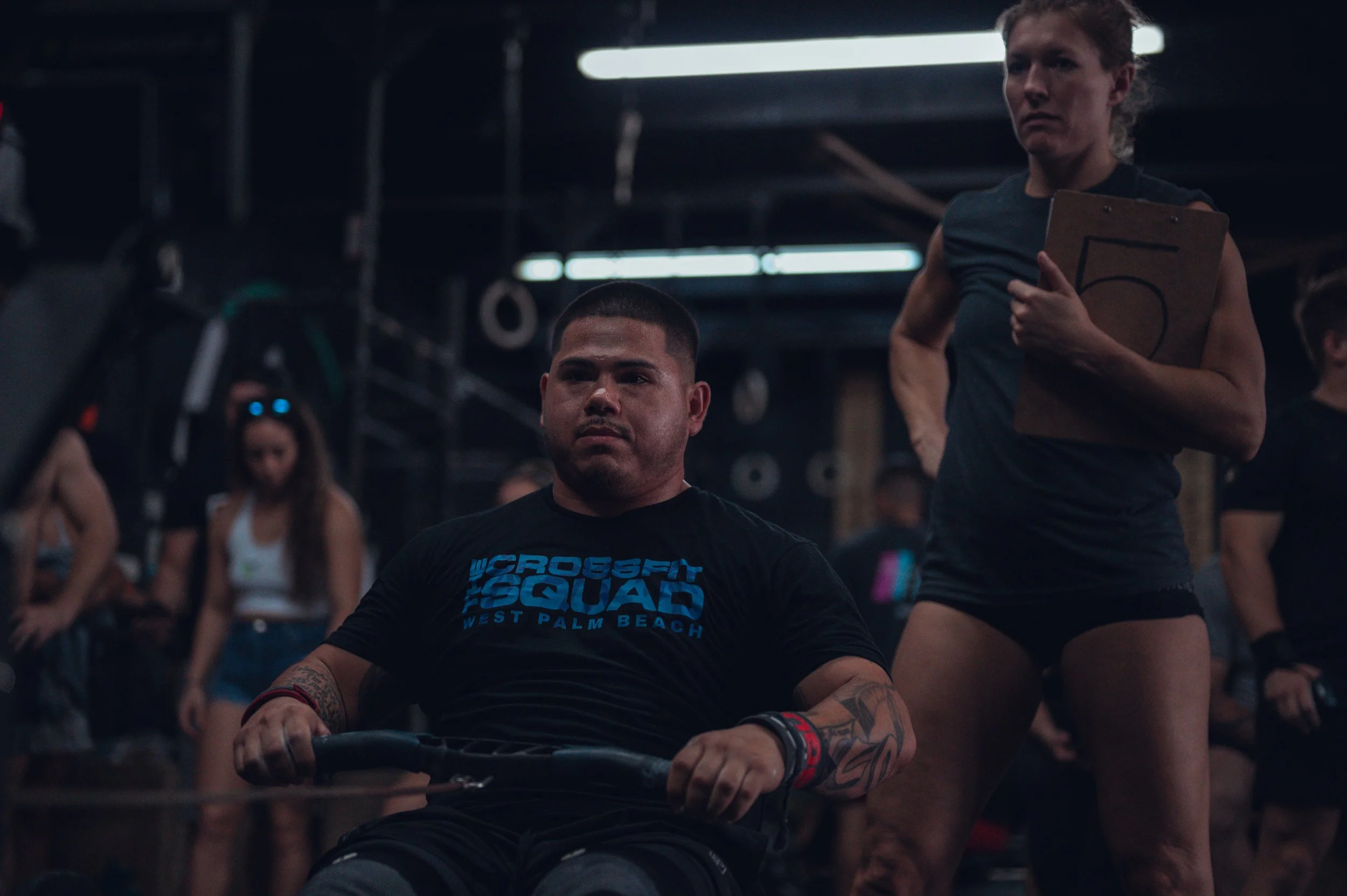 A man in a black CrossFit T-shirt sitting on a rowing machine, with a woman standing behind him holding a clipboard, in a gym filled with other people.