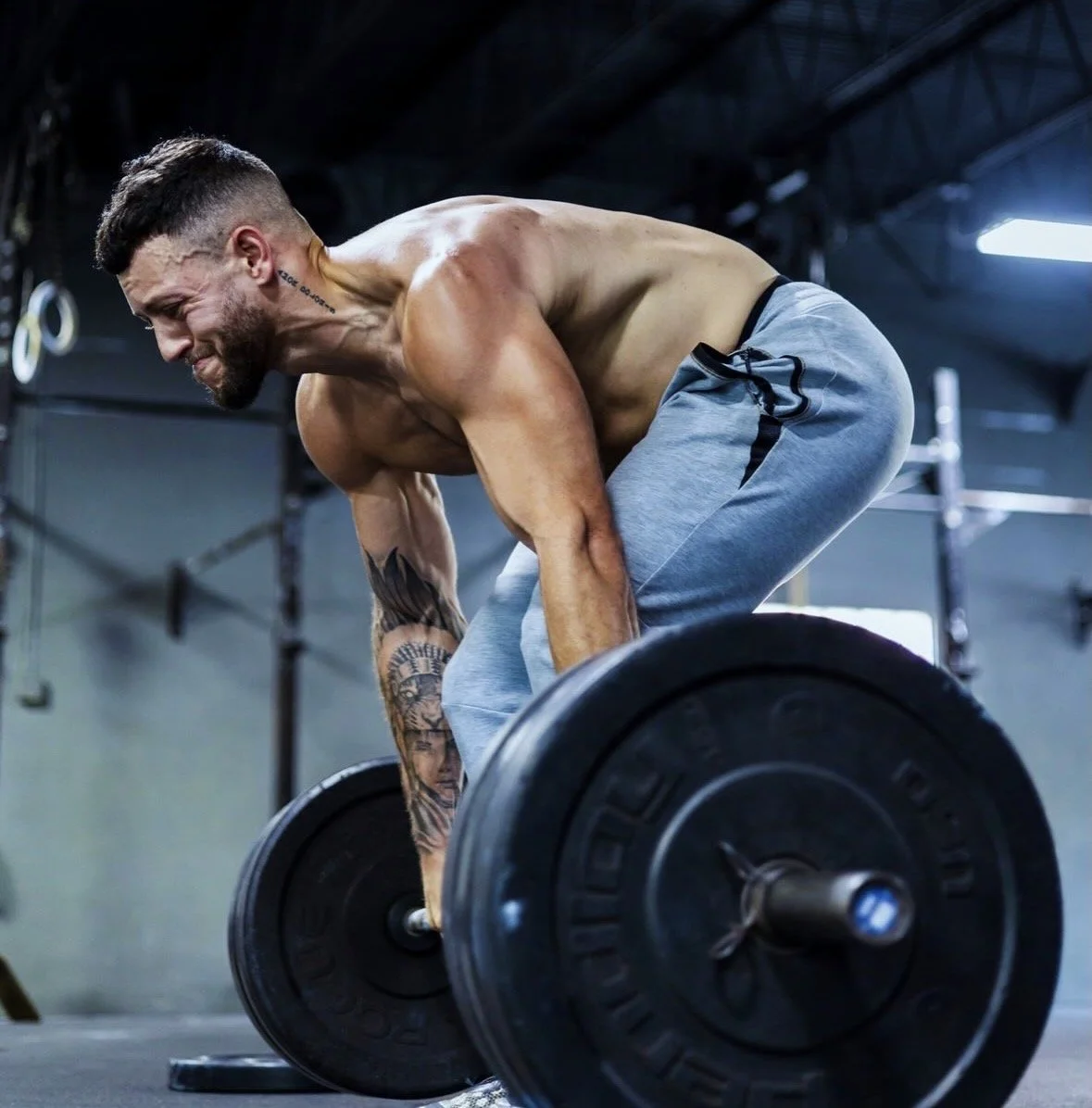 A man with tattoos lifts a barbell in a gym, wearing light gray sweatpants and shirtless, with focused effort.
