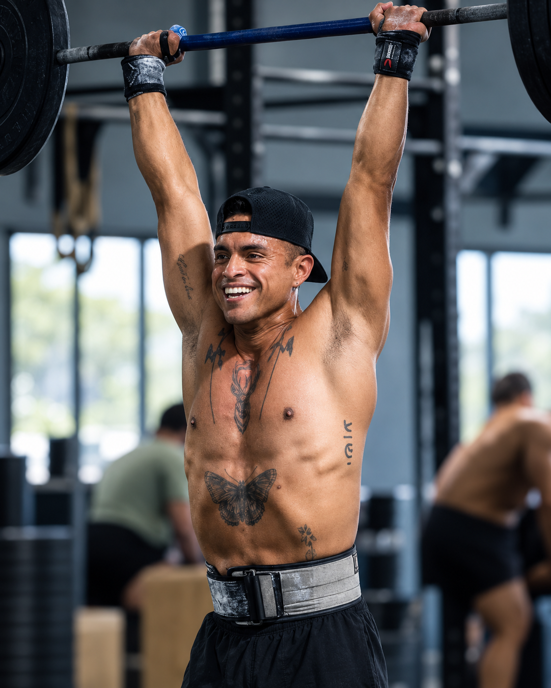 A shirtless man wearing a black cap and black workout shorts is lifting a barbell overhead in a gym, smiling. He has tattoos on his chest and arms, including butterflies and other designs.