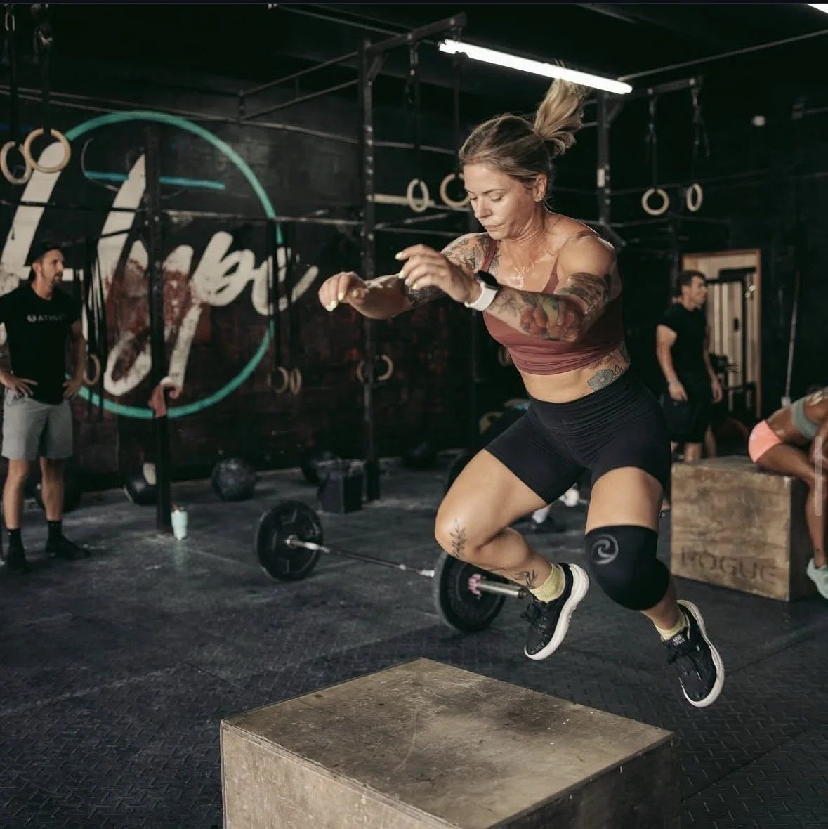Woman jumping onto a wooden box during a workout in a gym. Other people working out and gym equipment are visible in the background.