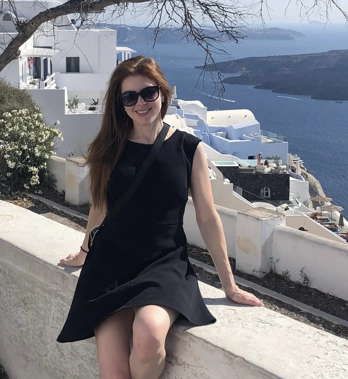 A woman wearing a black dress and sunglasses sitting on a white wall with a coastal village in the background, featuring white buildings and a large body of water.