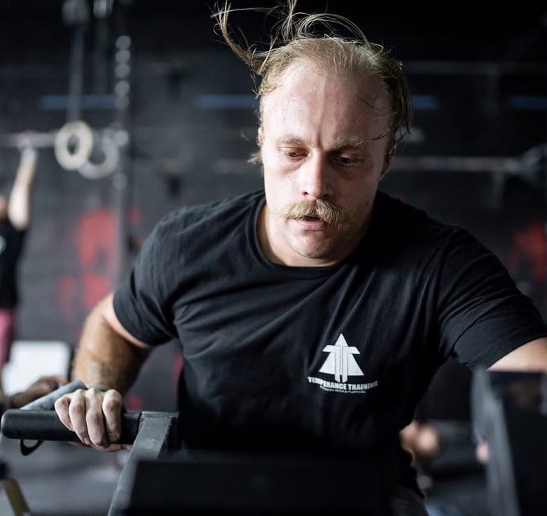 Man with a mustache and styled hair exercising in a gym, wearing a black T-shirt with a logo that says "Temperance Training," while holding a weight.