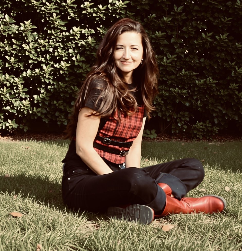 A woman with long brown hair sitting on the grass in front of a green hedge, wearing a black and red plaid top, black pants, and red boots, smiling at the camera.