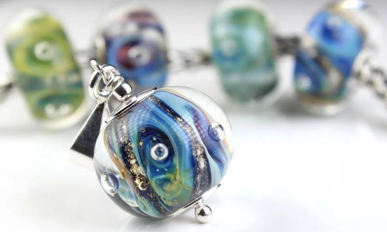 Close-up of a multicolored glass memorial bead on a silver chain with other similar beads blurred in the background.