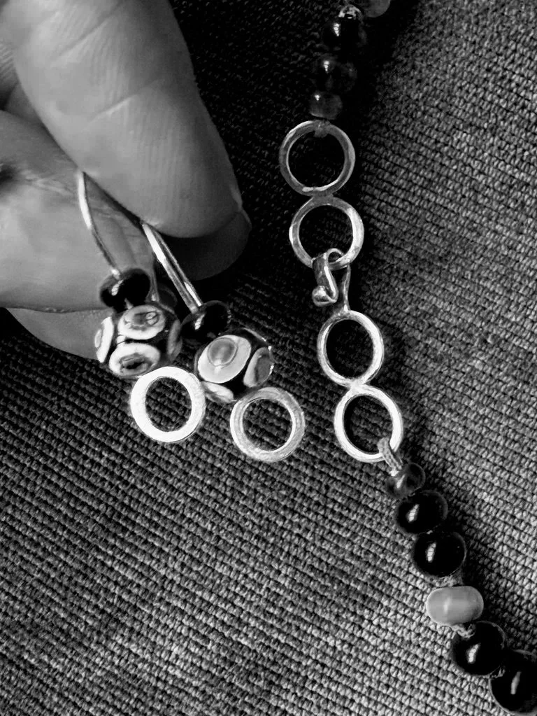 Close-up of a hand holding necklace with black and white beads and metal rings, resting on textured fabric.
