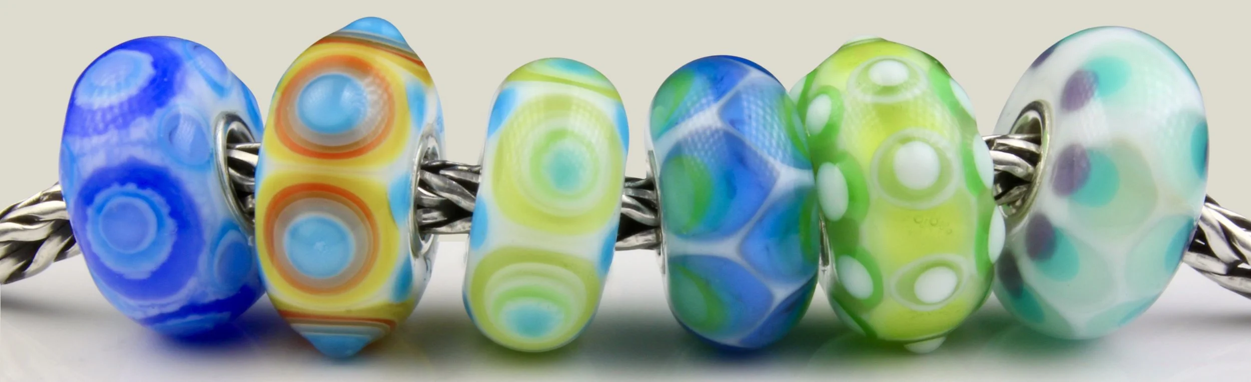 Close-up of six colorful glass beads threaded onto a foxtail chain, featuring bright swirling patterns in blue, yellow, green, and white.