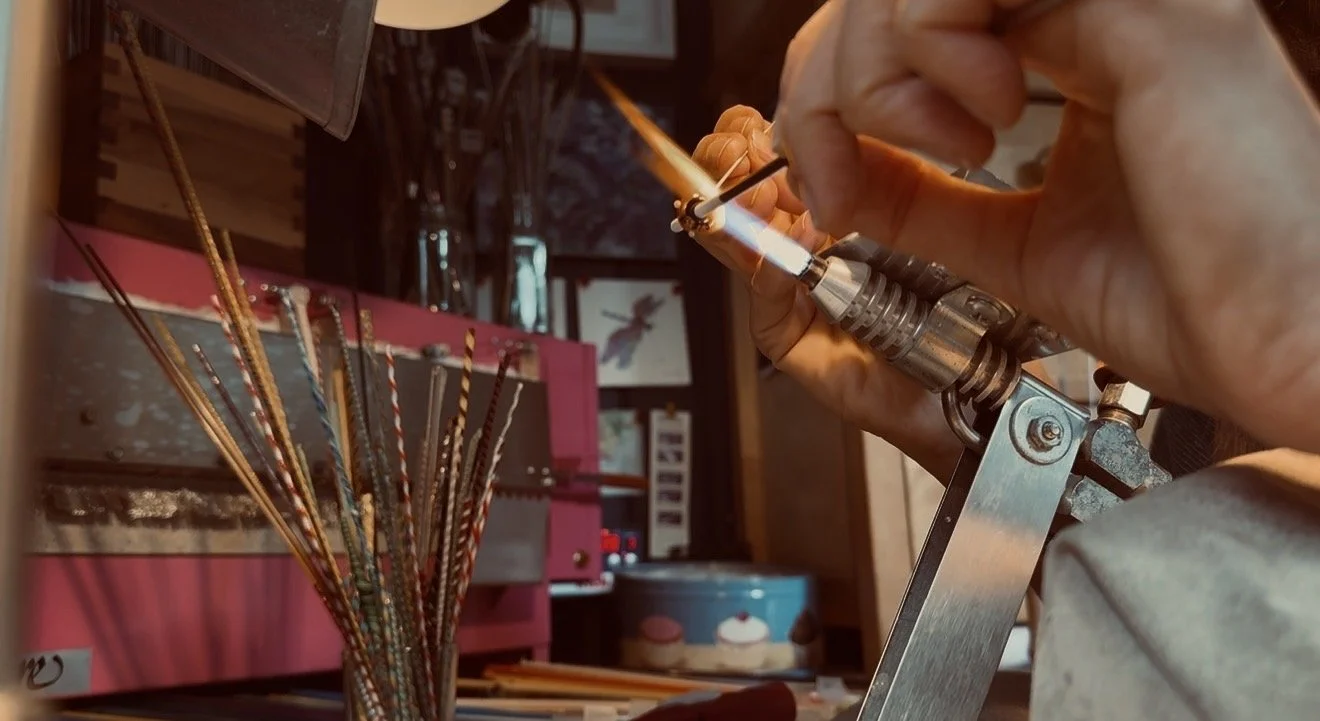 A person is using a torch to melt or solder a small object in a workshop or craft room, with a colorful assortment of sticks or wires in the background.