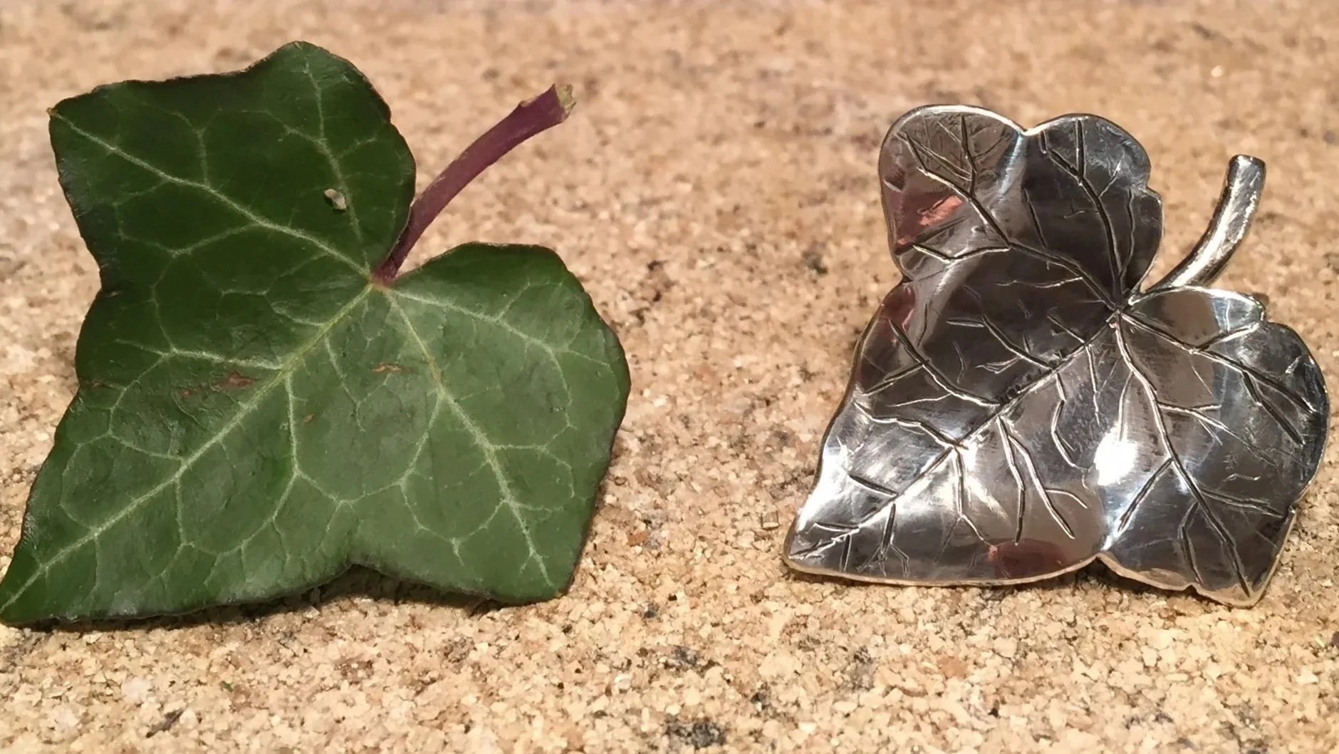 A green ivy leaf and a metal leaf-shaped object on a sandy surface.