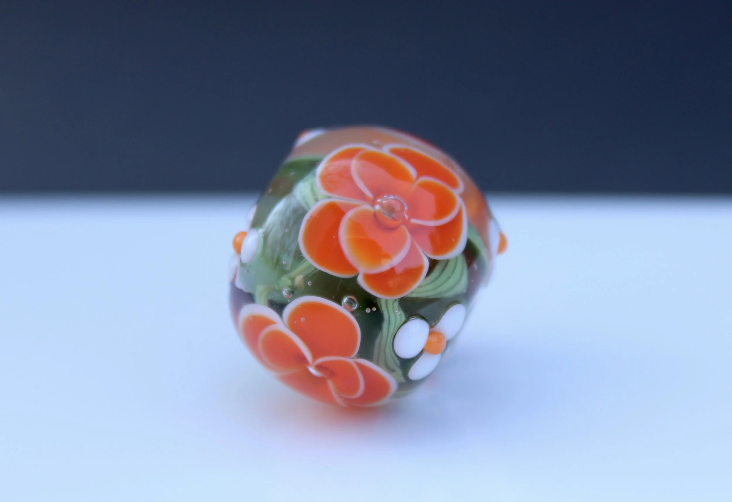 Close-up of a decorative glass marble with orange, white, and green floral patterns, reflected on a smooth surface under a dark background.