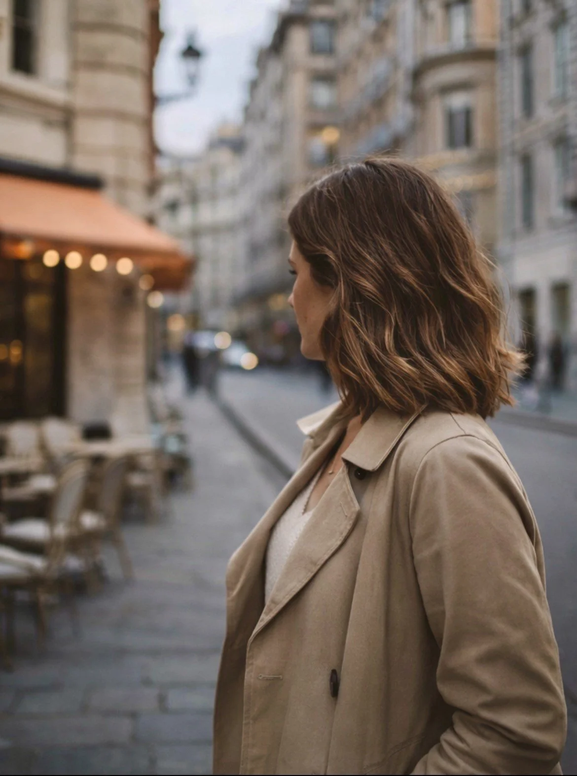 A woman with shoulder-length brown hair wearing a beige trench coat stands on a city sidewalk, facing sideways. Behind her, there are blurred buildings, outdoor seating, and a storefront with warm lights in an urban setting.