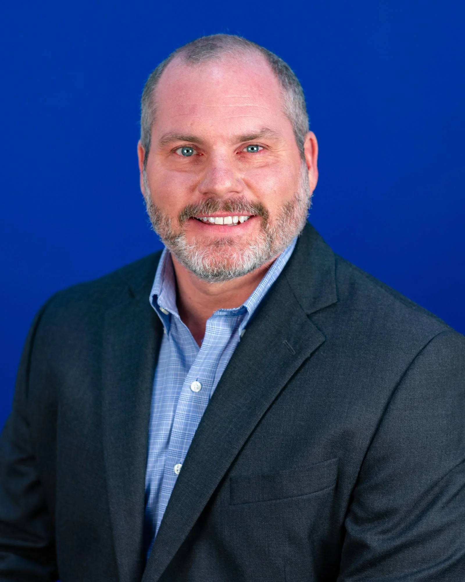 Close-up of a smiling middle-aged man with short gray hair, blue eyes, and a beard, wearing a dark blazer and a blue checkered shirt against a blue background.