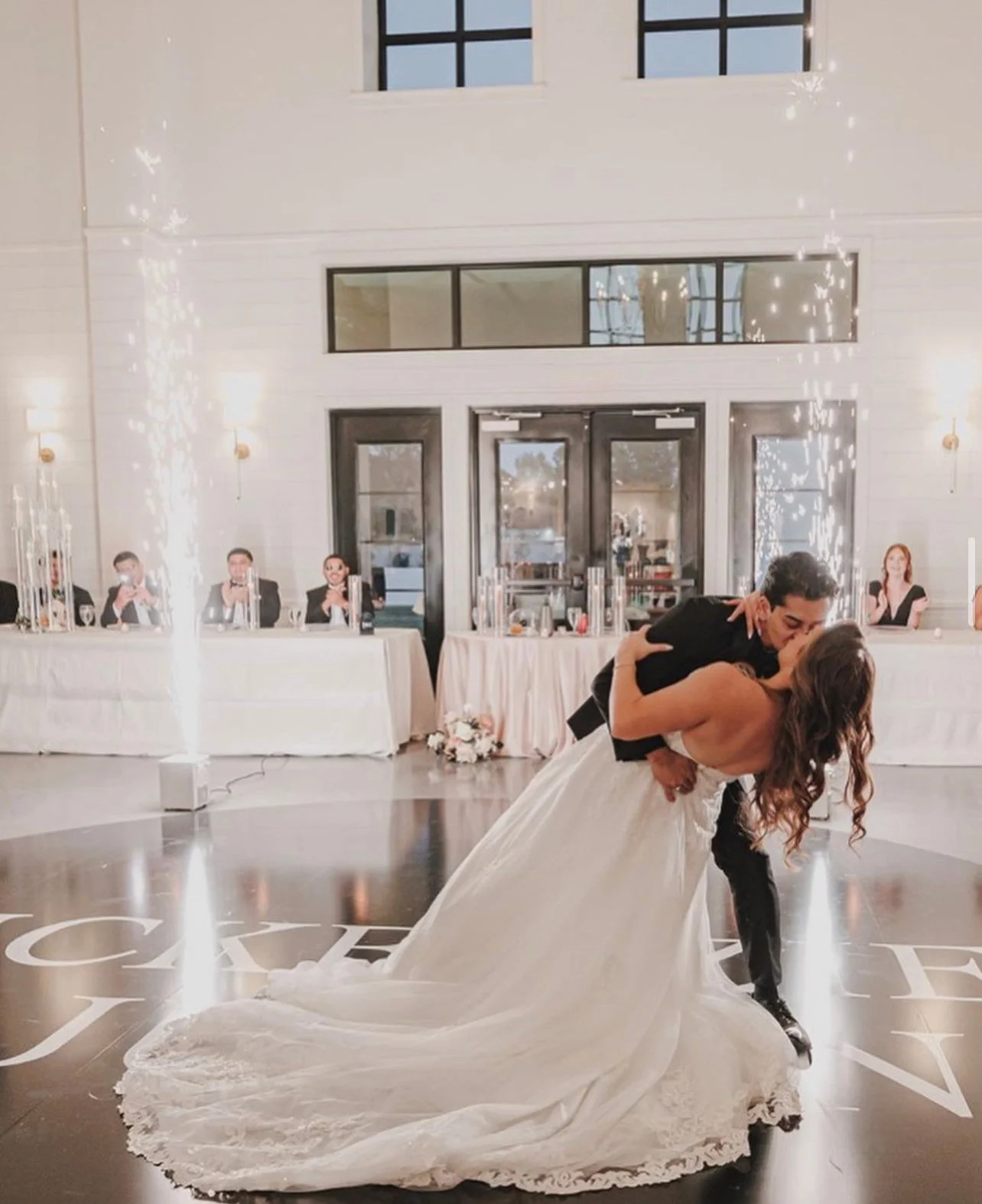 A bride and groom sharing a dance at their wedding reception, with the groom dipping the bride as they kiss. In the background, guests are seated at a long table, watching the couple. Sparkling lights are visible on the walls, and a bouquet is placed on the floor near the couple.