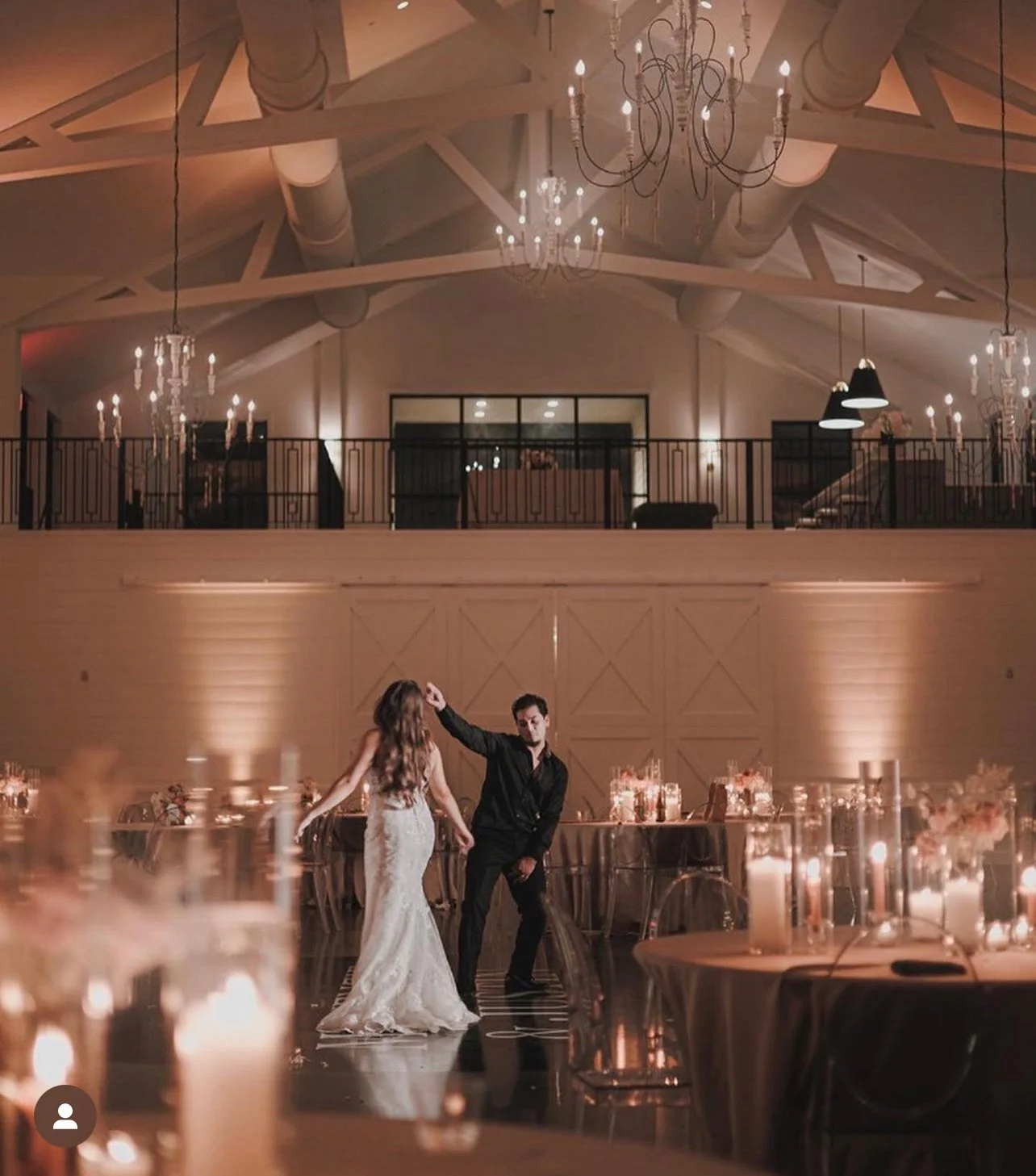 A couple dancing at a wedding reception in an elegant, warmly lit venue with high ceilings, chandeliers, and decorated tables.