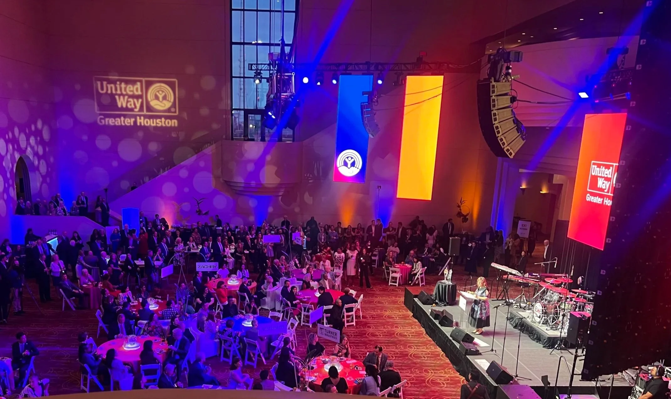 A large indoor event with many people seated at round tables, a stage with musical instruments, and large screens displaying logos for United Way Greater Houston, with purple and yellow lighting.