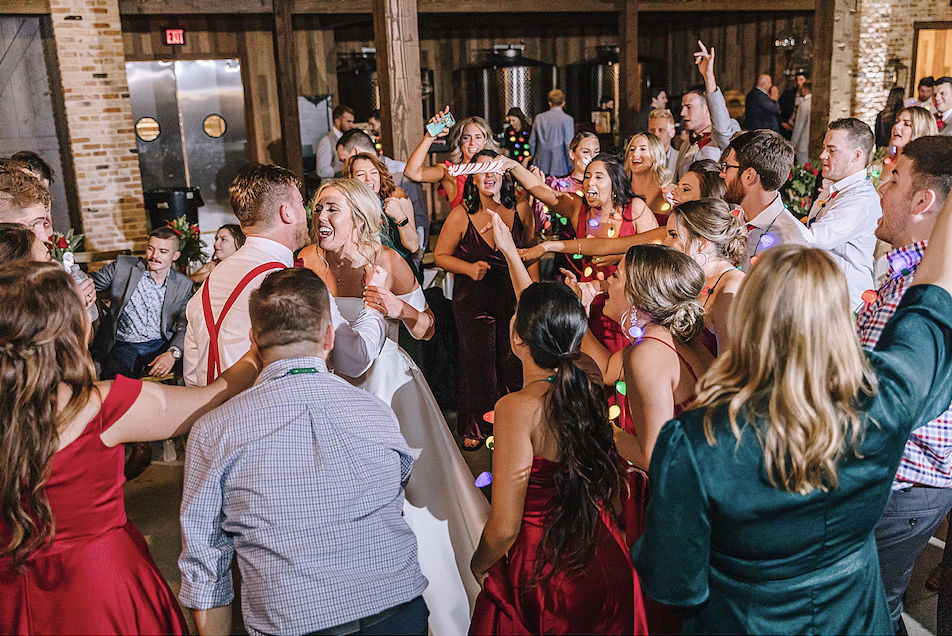 A group of people dancing and celebrating at a wedding reception, with some holding drinks and wearing colorful party accessories, in a rustic indoor venue.