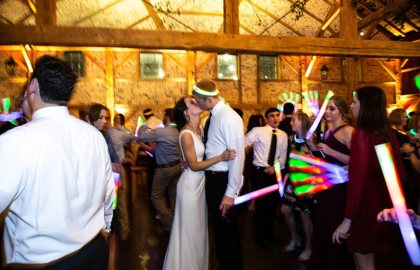 Couple kissing on dance floor at wedding reception, surrounded by guests with glow sticks in a rustic wooden venue.