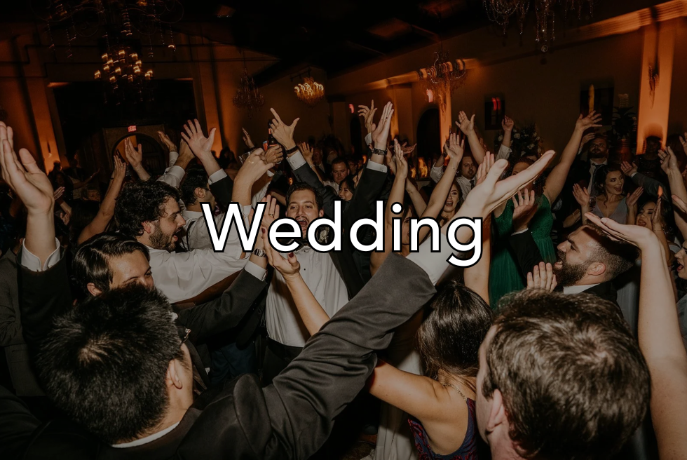 Crowd of wedding guests dancing and celebrating indoors with warm lighting and chandeliers.